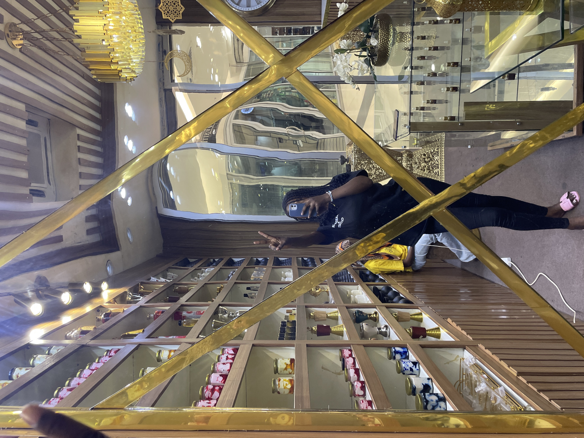 The image shows a person taking a selfie in a store that appears to be in Unguwar Sarki, Nigeria. The store has shelves filled with various items, including jars of what appear to be cosmetics or food products, and decorative items like vases and lanterns. The person is wearing a black t-shirt and black pants, and is holding a smartphone in their left hand. They are also making a peace sign with their right hand. The store has a modern design with a chandelier and wooden accents. The reflection in the mirror also shows part of a shopping mall.