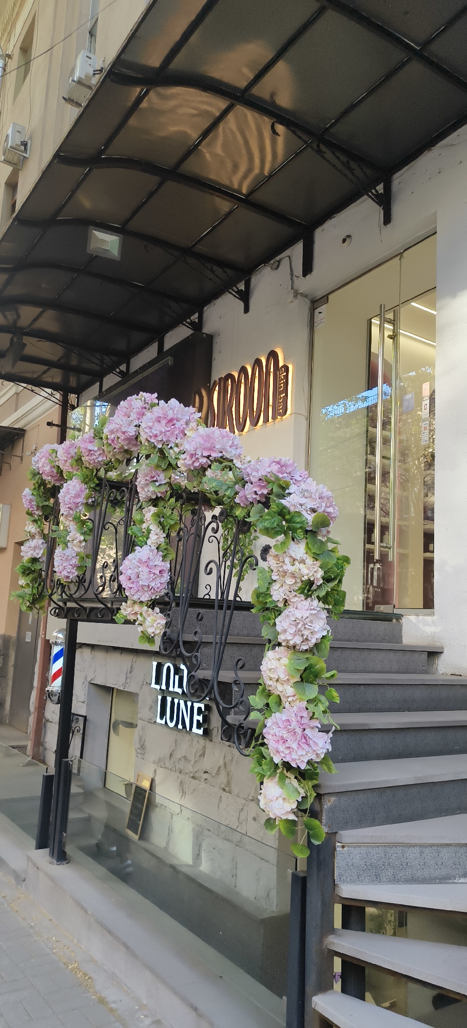 The image captures the exterior of a commercial building in Yerevan, Armenia, featuring architectural and decorative elements. The building has a dark-framed canopy extending over the entrance, with air conditioning units visible on the upper facade. The entrance is marked by a sign reading "SKINROOM" with "skin bar" in smaller letters, illuminated in a warm tone. A set of gray concrete steps leads up to a glass-enclosed doorway.
The dark metal railing that borders the steps is adorned with an elaborate arrangement of pink and white flowers and green foliage. A sign below the railing reads "LUO LUNE" in capitalized letters. A traditional barber pole is positioned on the left of the railing.
The scene appears to be street-level, with portions of the sidewalk and street visible. The photograph is taken in daylight, with moderate lighting.