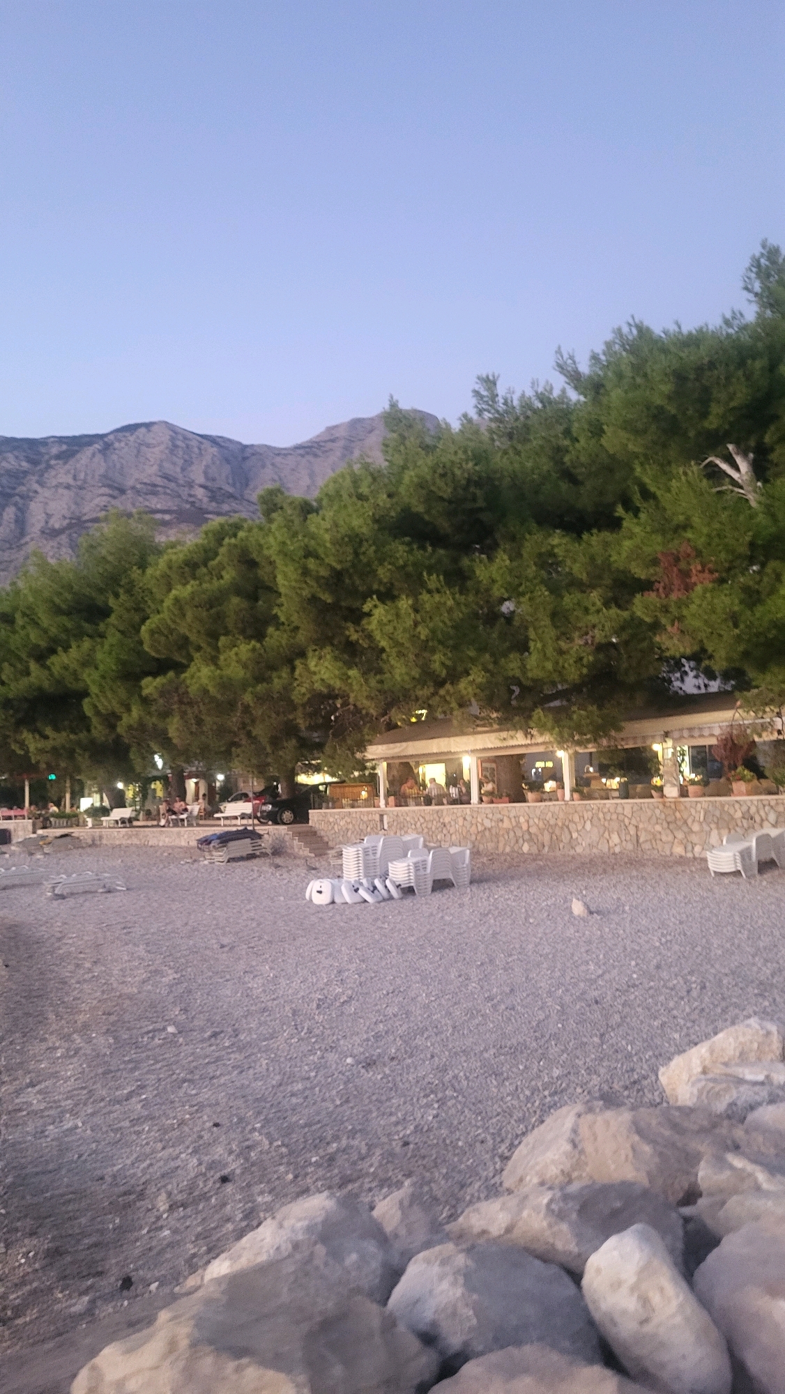 The image depicts a beachside scene in Općina Tučepi, Croatia, during twilight. In the foreground, large rocks are scattered on a pebble beach. Further back, rows of white plastic beach chairs and lounge chairs are neatly arranged, some stacked. A pathway separates the beach from a stone-walled embankment lined with potted plants. Behind the embankment, a building with an awning and outdoor seating is visible, illuminated by lights. Several people are seated at tables inside and outside the establishment. A black car is parked near the building. A dense line of green pine trees forms a backdrop to the building, and in the far distance, a mountain range is silhouetted against the pale sky.