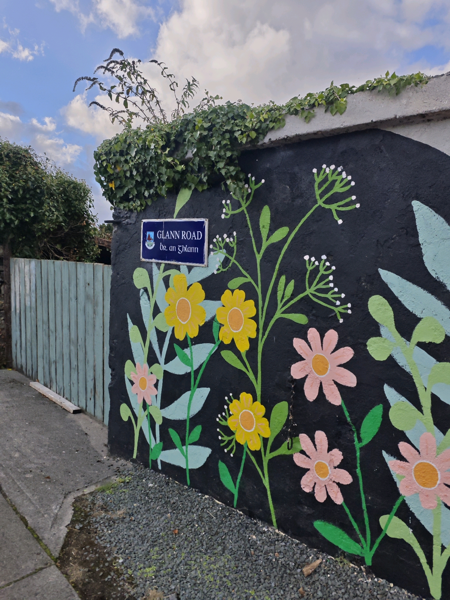 The image depicts a section of a mural painted on a black wall, likely an exterior wall in Oughterard, Ireland. The mural features stylized flowers and foliage in vibrant colors, including yellow, pink, and light green, against the dark background. A blue street sign with white lettering, reading "GLANN ROAD" and "br an Ghleann," is affixed to the wall, indicating the location. To the left of the mural is a weathered, light-blue wooden fence. The ground in front of the wall is a mix of pavement and dark gravel. Overhanging the top of the wall is dense green ivy, and above that, a blue sky with scattered white clouds. The overall scene is one of urban street art integrated with natural elements.