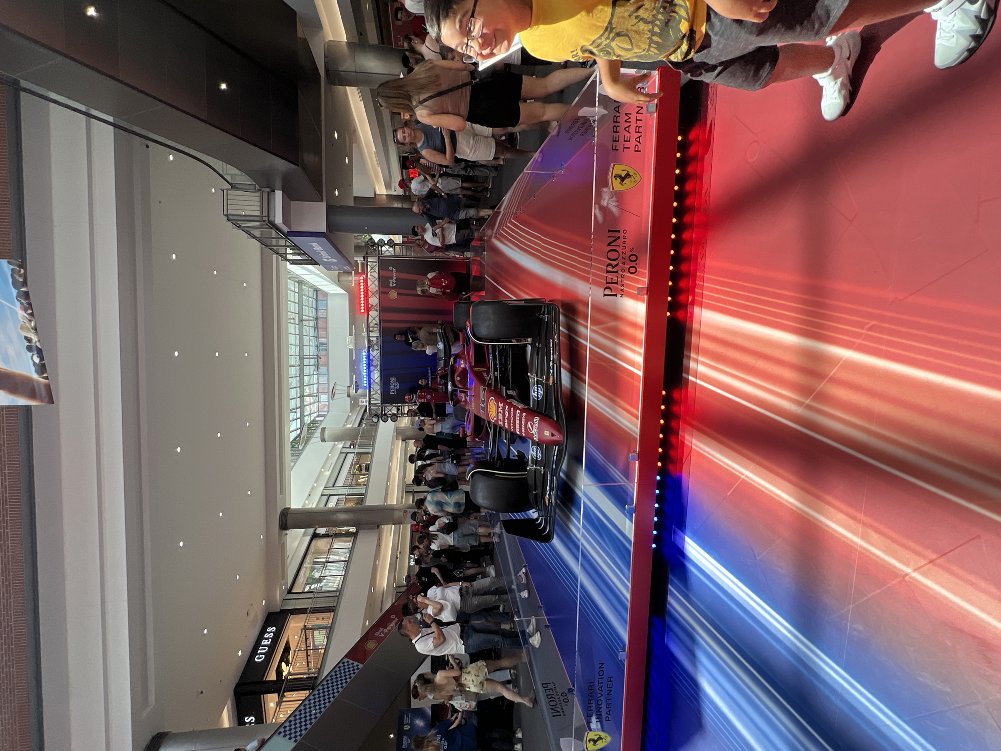 This image depicts a Formula 1 racing car on display inside a shopping mall. The car, which is red and prominently branded with "Peroni" and "Ferrari Team Partner," is positioned on a platform illuminated with red and blue streaks of light. A crowd of people, dressed in casual summer clothing, are gathered around, observing the display. Some are taking photos, while others are engaged in conversation. In the background, the typical architecture of a shopping mall can be seen, including storefronts like "Guess" and escalators. A young boy in a yellow shirt and glasses is in the foreground to the right, looking towards the camera and smiling. The overall atmosphere appears to be one of public engagement and excitement around the F1 display. This event appears to be taking place in Budapest, Hungary, as indicated by the presence of the Peroni beer brand, a known sponsor of Formula 1.