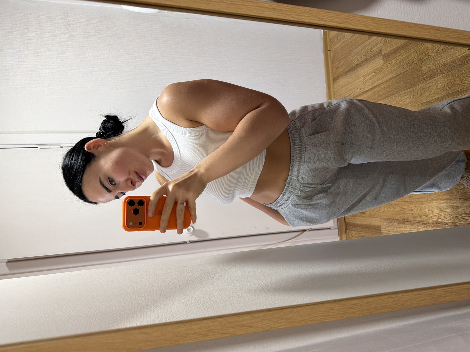 A young woman is captured in a mirror selfie, seen from the waist up, as she photographs herself using an orange smartphone. The image is oriented sideways, with the woman appearing to lean down towards her phone. She has dark hair styled in a messy bun and a small silver stud earring in her visible left ear. She is wearing a fitted white ribbed crop top that exposes her midriff and light grey sweatpants with gathered ankles. A silver ring is visible on her left ring finger as she holds the phone.
The mirror she is using has a light wooden frame and reflects a bright, minimalist indoor setting. Behind her in the reflection, there is a plain white wall, possibly a door or wall paneling, and a light-colored wooden floor. The overall lighting appears soft and even, suggesting an indoor environment without harsh shadows. Her expression is neutral as she looks down at the phone screen, focused on taking the picture. No visible text is present in the image.