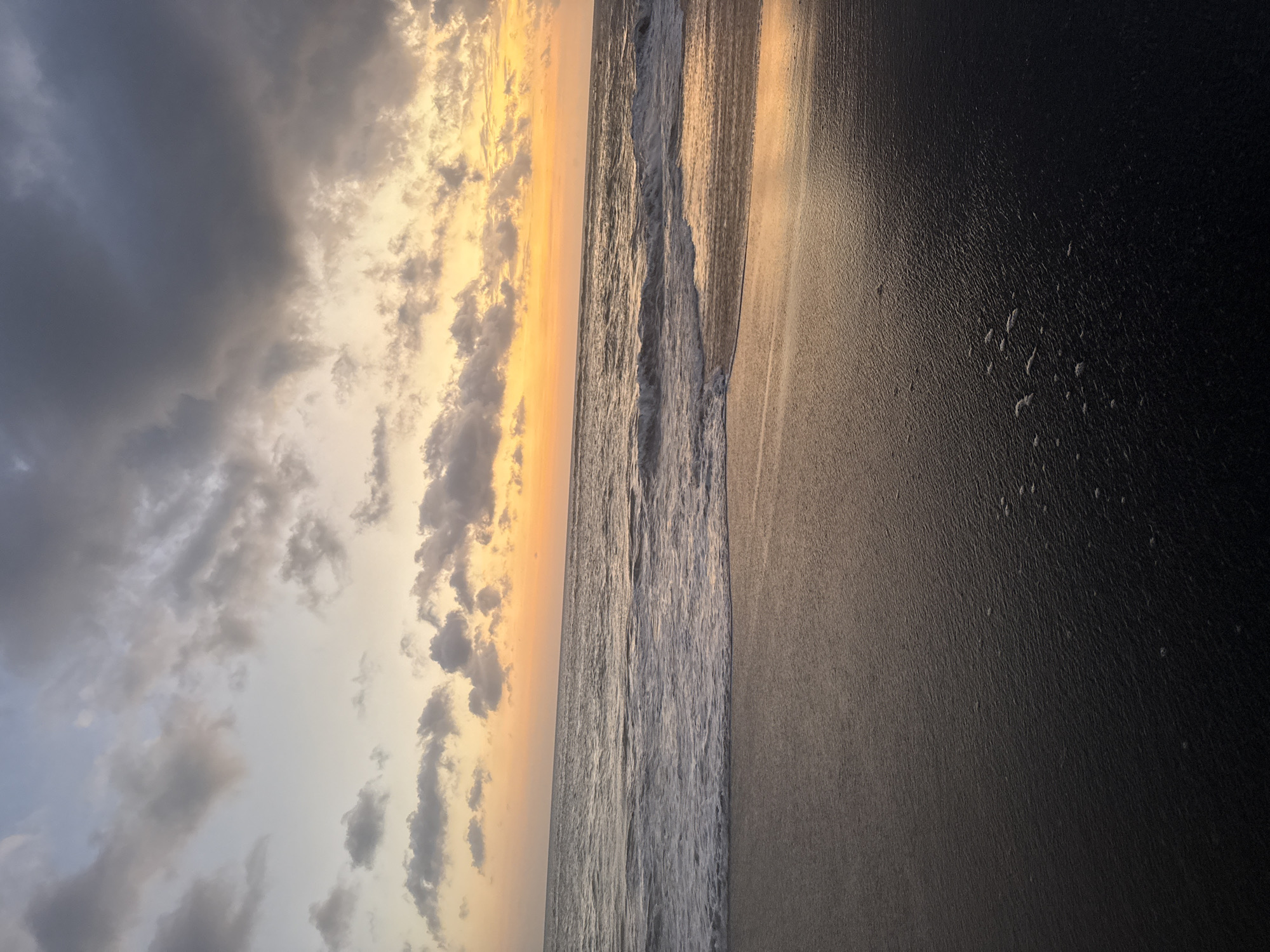 The image depicts a beach scene at sunset. The sky is filled with clouds, with hues of gray and yellow near the horizon. The ocean waves are visible, and the sand is dark. The location is Unknown, Unknown.