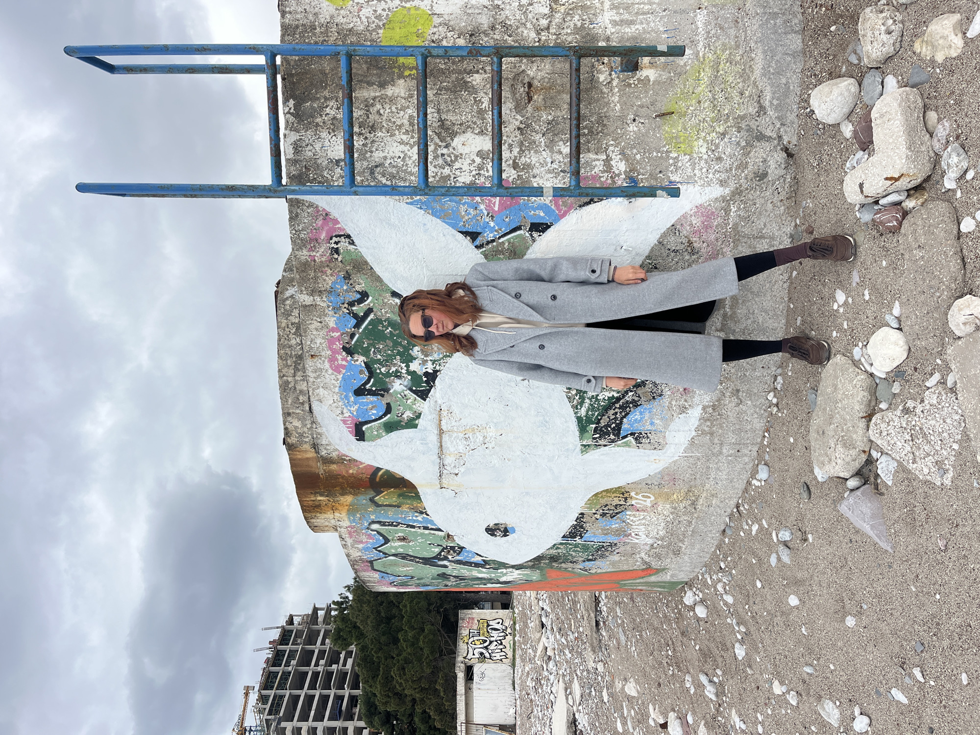 A woman in a grey coat and sunglasses poses against a concrete structure with a painted dove and graffiti, located on a pebble-strewn area. A rusty blue metal ladder is attached to the concrete structure. In the background, there is an unfinished building, trees, and a cloudy sky. The woman is wearing brown shoes, black leggings, and a light-colored top under the coat. Based on the presence of the pebble beach and the building style, the location is likely in or near Budva, Montenegro.