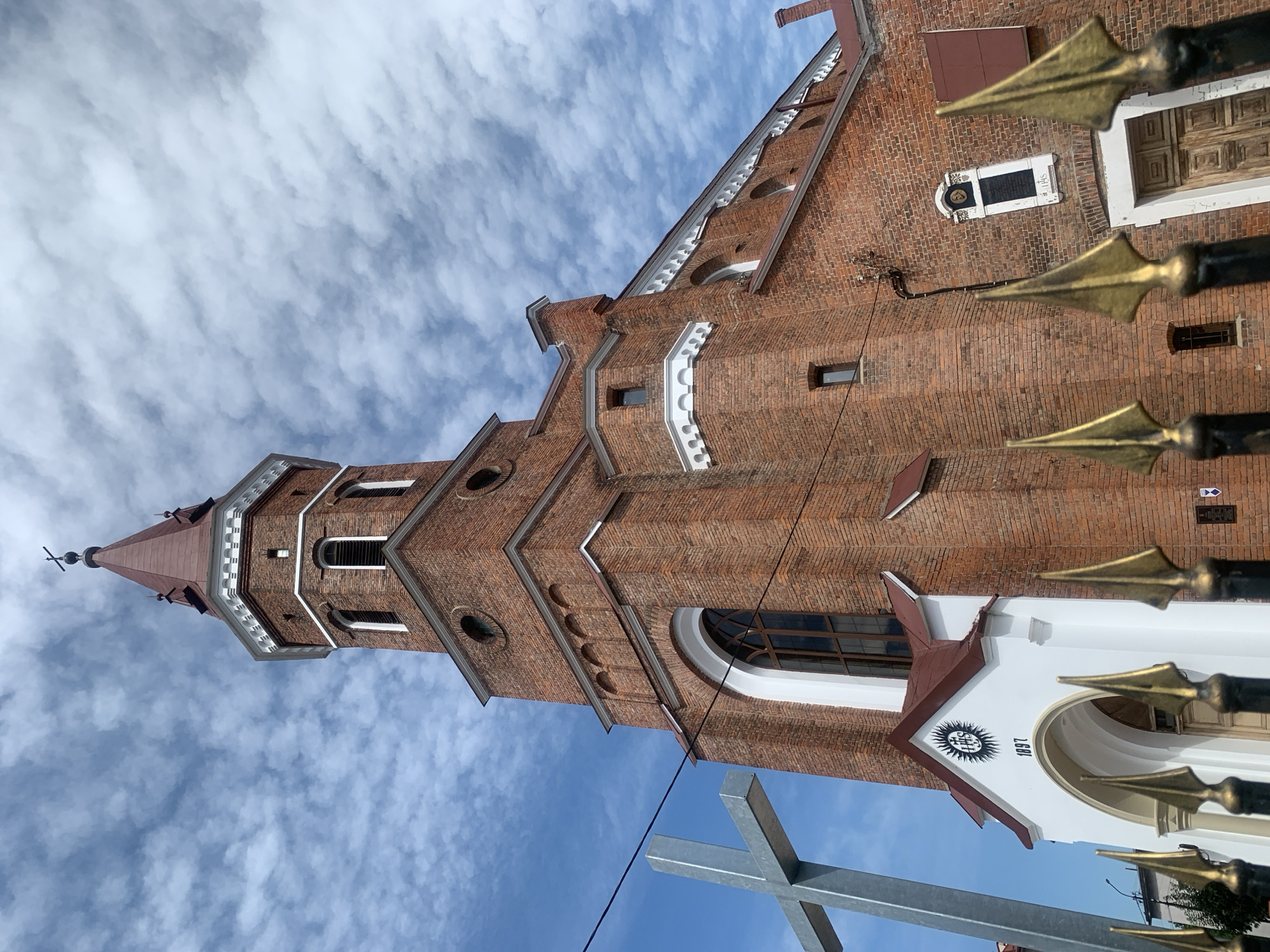 The image shows a brick church with a tall tower against a bright blue sky with white, wispy clouds. In the foreground, a large metal cross is visible on the left side. A black metal fence with gold-colored finials runs across the bottom of the image. The church has arched windows and decorative white trim. Above the main entrance, there is a white facade with a sun symbol and the year "1897" inscribed. The church is located in Kolomyia, Ukraine. The image is taken from a low angle, looking up at the church tower.