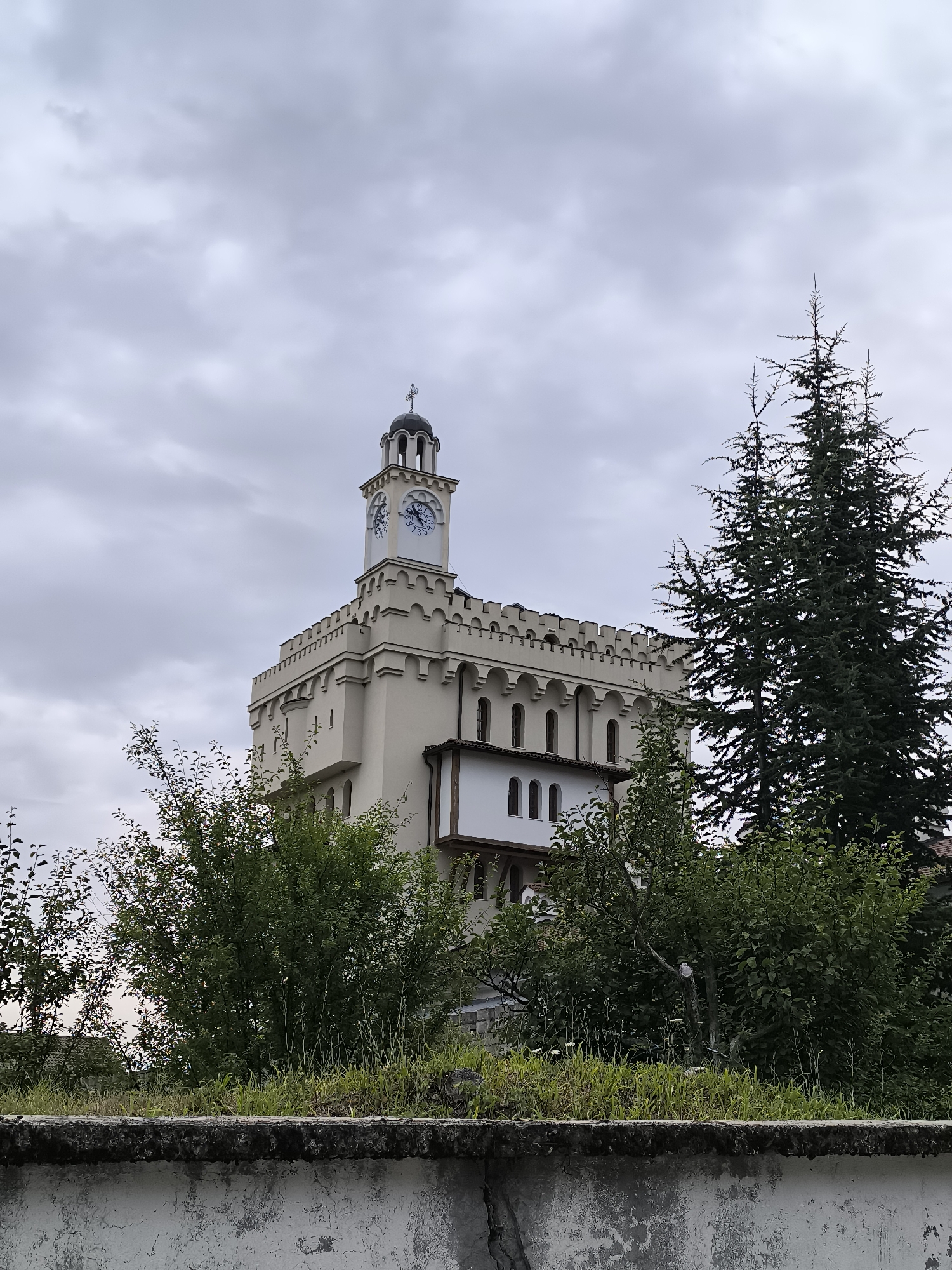 The image captures a view of the Church of St. George in Hadzhidimovo, Bulgaria. The church, built in the style of a fortress, features a prominent bell tower with a clock face, topped with a dome and a cross. The building is constructed of light-colored stone and has crenellated battlements along its upper edge. Green trees and foliage partially obscure the lower portion of the structure. In the foreground, a weathered concrete wall runs horizontally across the bottom of the frame. The sky is overcast with gray clouds.