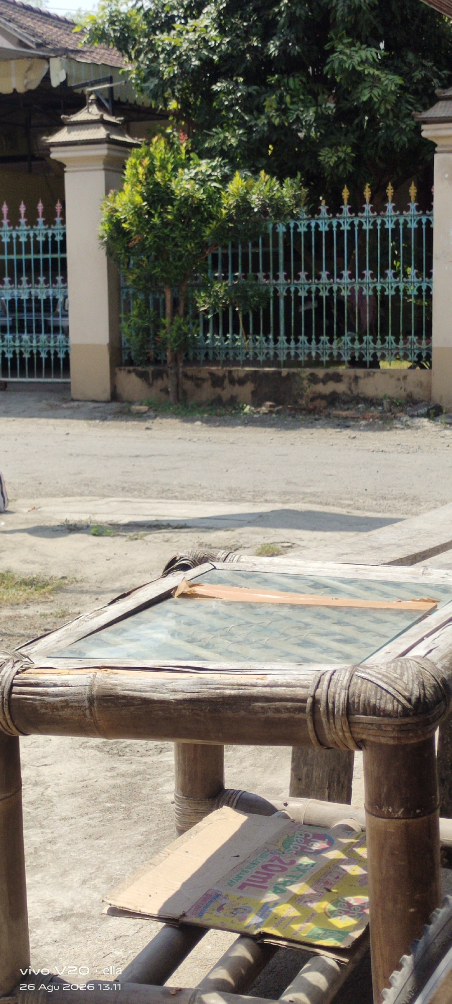 This is a vertical, outdoor, eye-level, full shot of a bamboo table with a glass top, positioned in the foreground. The table is weathered and appears to be handmade. A piece of cardboard is resting on the table below the glass top. In the background, a turquoise metal fence with decorative finials runs horizontally across the image, separating the foreground from a paved area and a building. The building has a tiled roof and a pillar with a decorative element. Several trees and shrubs are visible behind the fence. The ground appears to be a mix of dirt and gravel. The lighting suggests it is daytime, with shadows cast on the ground. A watermark in the bottom left corner indicates the photo was taken with a "vivo V20" phone on "26 Agu 2026 13.11" and the location is "Simo, Indonesia". No people are visible in the frame.
