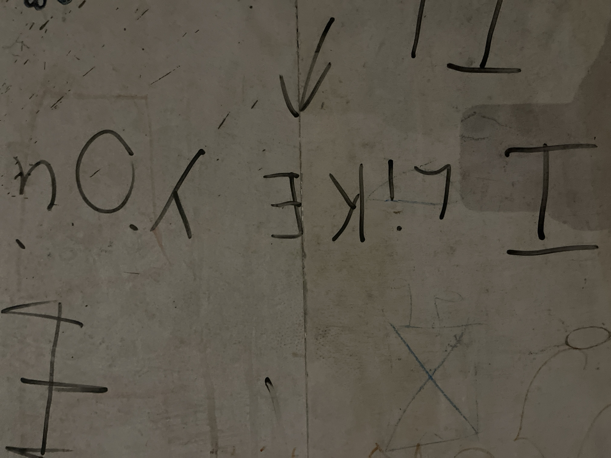 The image is a close-up, eye-level, medium shot of a dirty, off-white wall with graffiti. The graffiti consists of handwritten text and drawings in black and blue. The main text reads "I LIKE YOU" in large, blocky letters. To the left of the text, there's a large, stylized "I" and a drawing that appears to be a kite or a diamond shape with an "X" inside, drawn in blue. To the right of the text, there are some additional drawings, including what looks like a capital "I" with horizontal lines, and what could be the beginning of another word. There is also an arrow pointing upwards. The wall itself is textured and appears to be stained and marked in various places, suggesting it is in a public or neglected area. The lighting is dim and uneven, casting shadows on the wall.
