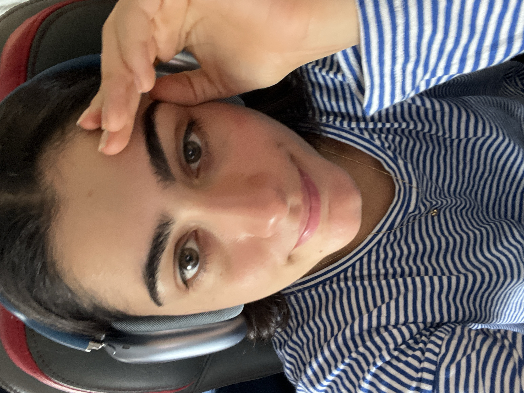 A close-up, high-angle, selfie-style shot features a young woman in Paray-Vieille-Poste, France. She is wearing blue and white striped long-sleeved shirt and large, light blue over-ear headphones. Her right hand is raised and touching her forehead and temple, obscuring the right side of her face. Her left eye is visible and looking directly at the camera. The background consists of the dark grey and red upholstery of a chair.