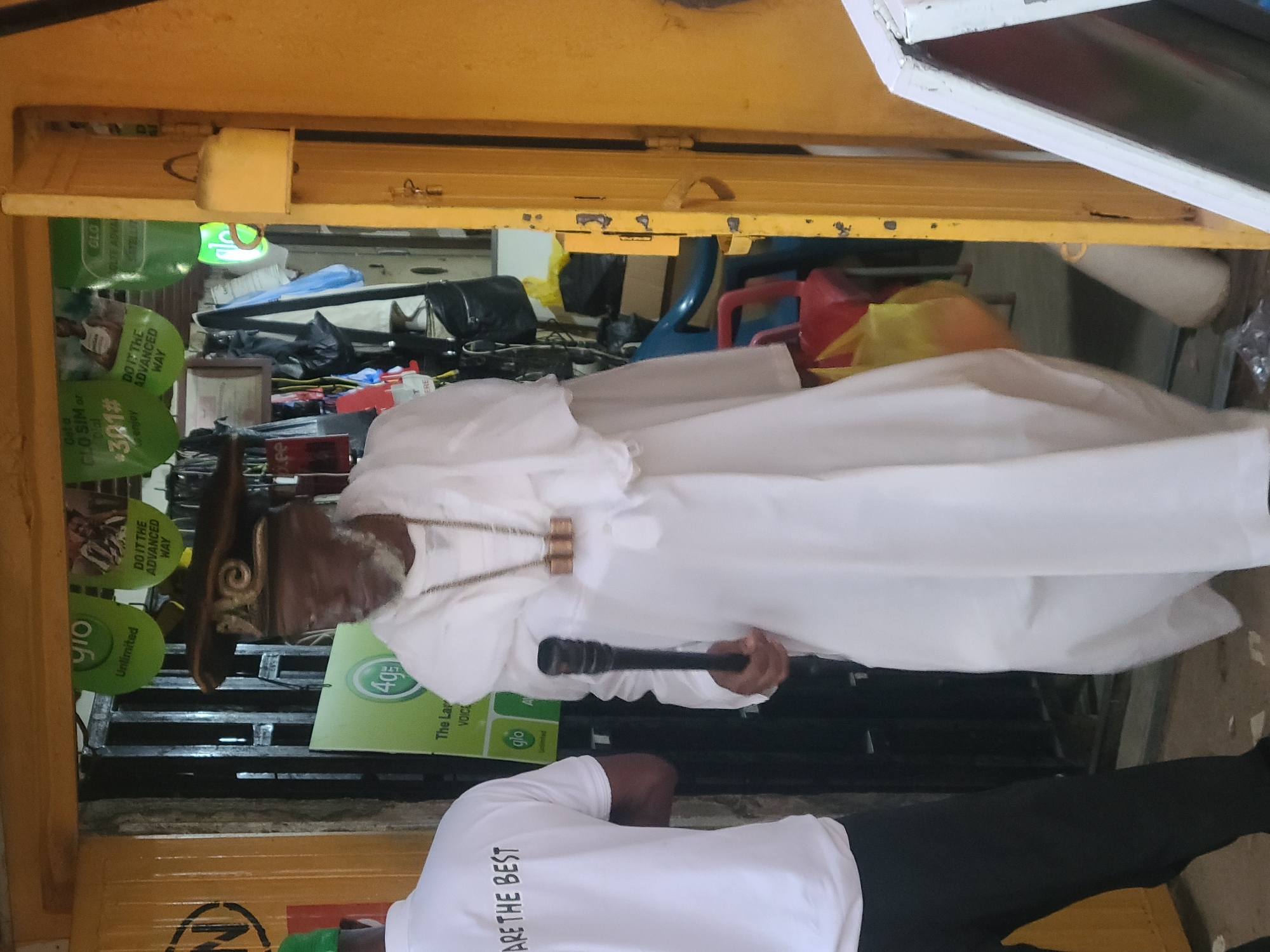 An outdoor, eye-level, full-shot image captures a man in traditional white attire walking through a yellow doorway in Jos, Nigeria. He is adorned with a unique wooden hat and layered necklaces, and he carries a black staff. The man is partially obscured by a second individual wearing a white polo shirt with black lettering that reads "ARE THE BEST" and black pants, who is standing to his left. Behind the main subjects, a shop interior is visible, featuring shelves stocked with various items, including green promotional banners with the "Glo" logo, suggesting a telecommunications store. The doorway frames a bustling urban environment with hints of activity beyond.
