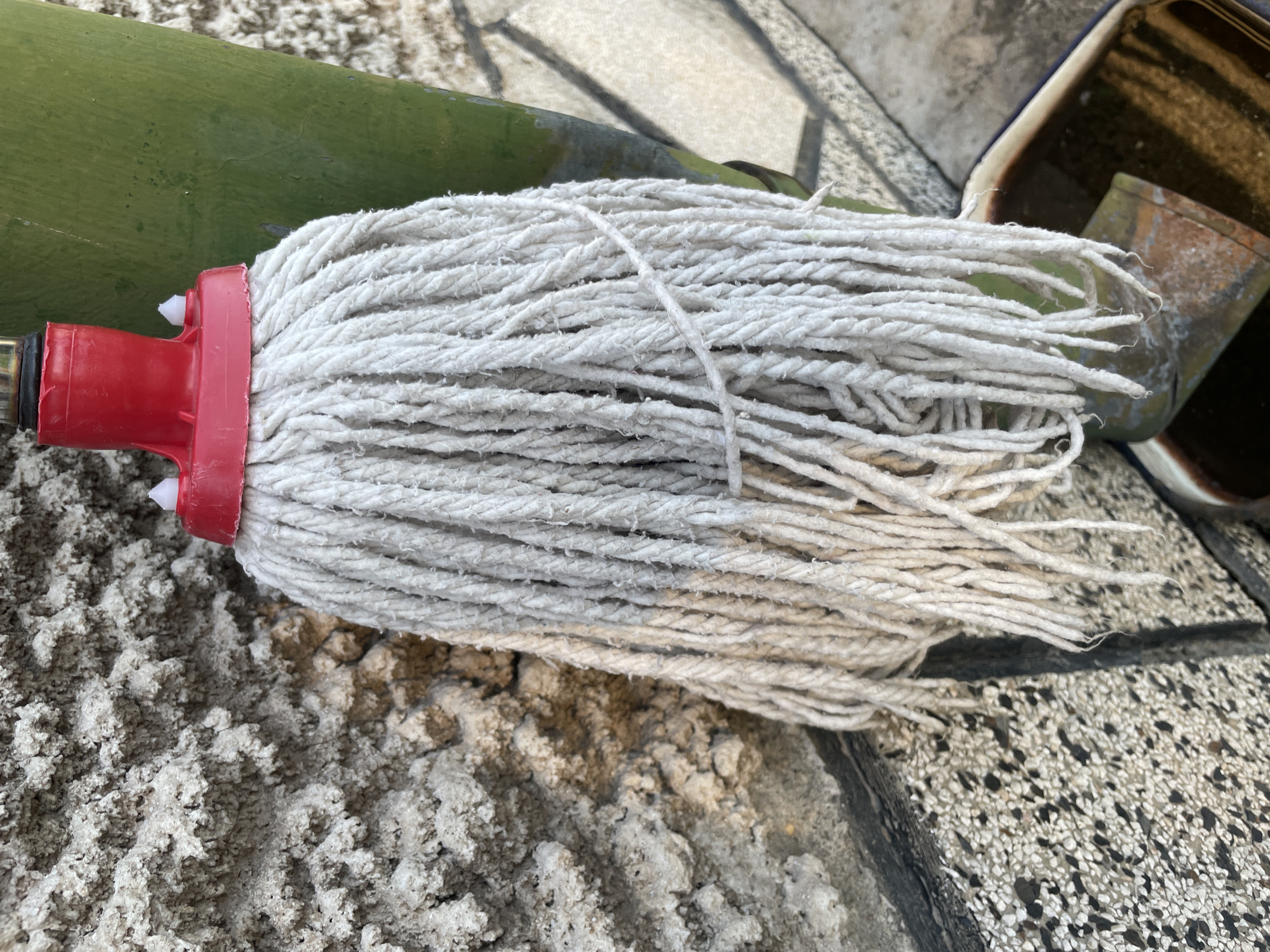 The image shows a cotton string mop head with a red plastic connector lying on a textured, light-colored surface. A section of a green bamboo pole is visible adjacent to the mop. The edge of a container, possibly a bucket or pail, is also visible. The location is undetermined and there is no evidence to suggest that it is in Sofia, Bulgaria. There are no people in the image.