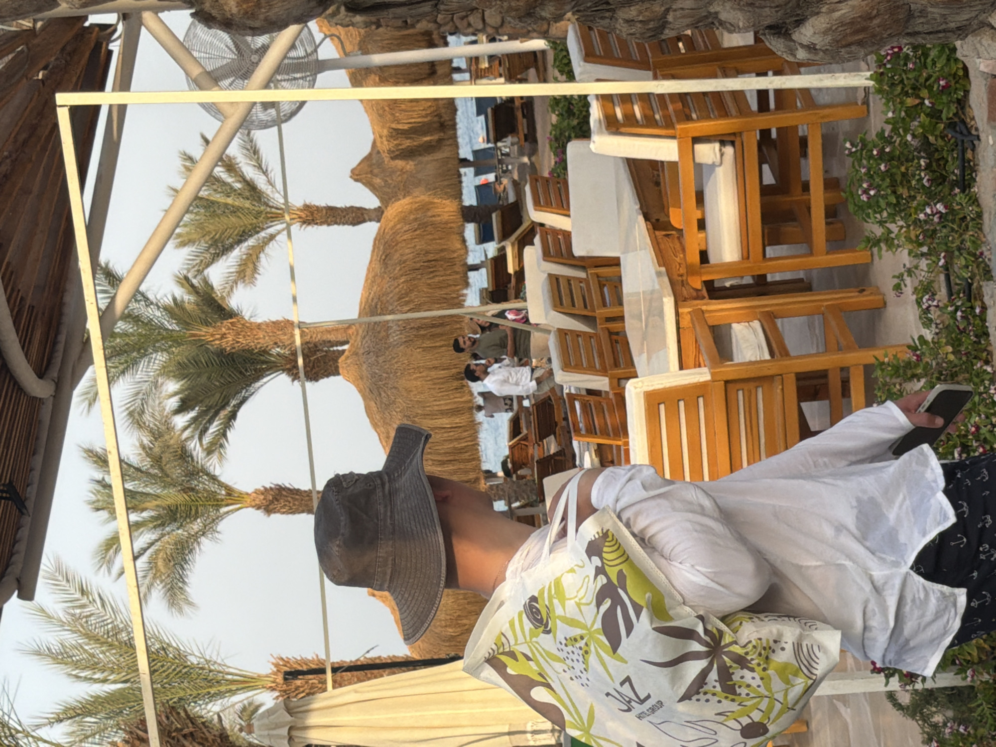 A man wearing a wide-brimmed hat and a white shirt is seen from behind, carrying a patterned tote bag over his left shoulder. He is standing in an outdoor seating area with wooden tables and chairs. In the background, palm trees and thatched umbrellas are visible, suggesting a resort or beach setting in Sharm El Sheikh, Egypt. The ocean can be seen in the distance. Other people are present in the background, some walking and others seated. The lighting suggests it is either early morning or late afternoon.