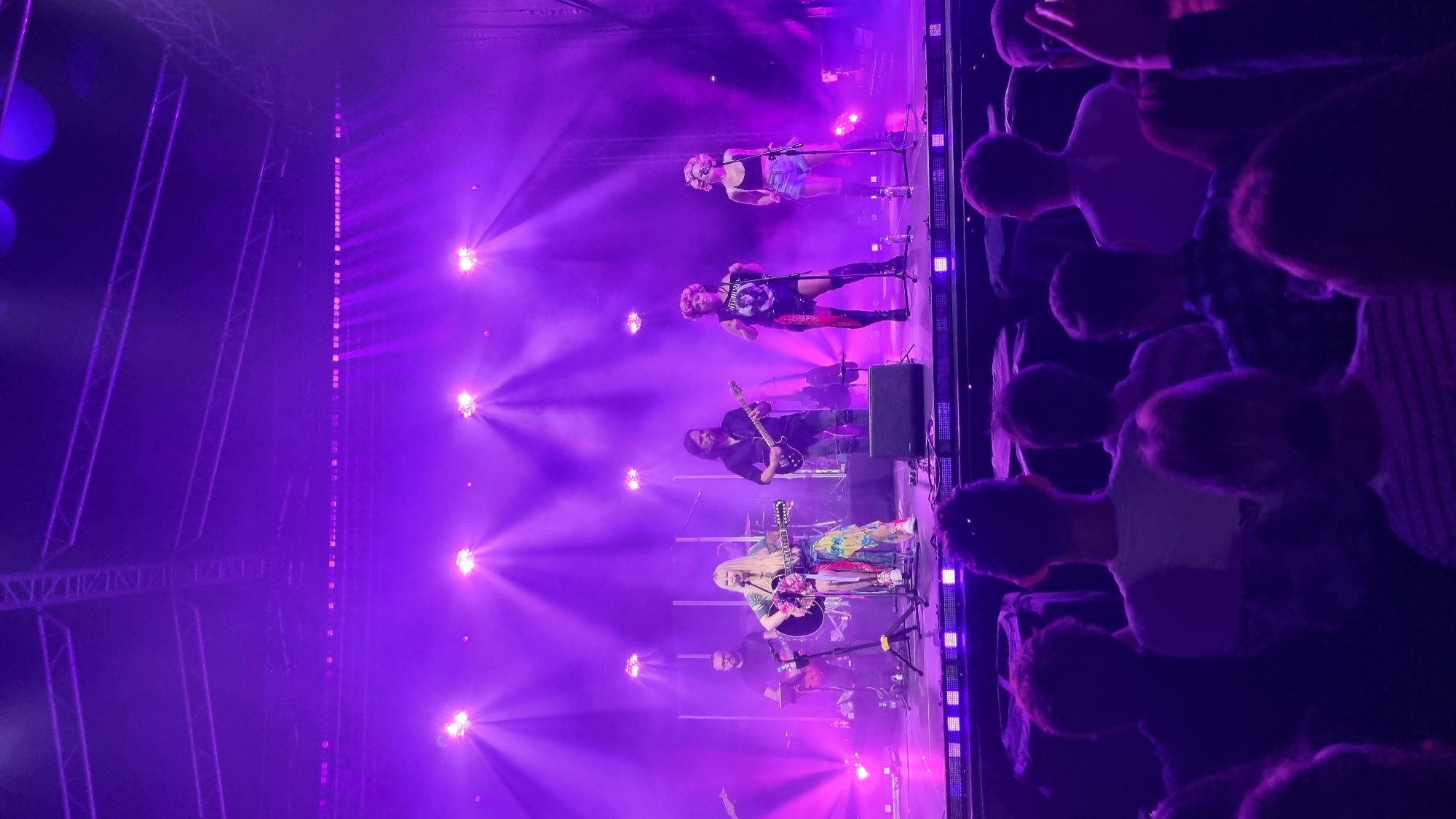 On a stage bathed in purple light, a band performs for an audience. The lead singer, a woman with long blonde hair, wears a colorful, flowing dress and plays a guitar. To her left, a man in a black shirt plays a guitar, and to her right, two women in dark tops and shorts, one with curly blonde hair, stand near microphones. The stage is equipped with multiple spotlights and speakers. In the foreground, members of the audience, mostly silhouetted, are visible from behind, facing the stage. The overall atmosphere is that of a live concert. The image appears to have been taken at the Municipal Sports and Recreation Center in Pruszcz Gdański, Poland, based on the context of a concert event.