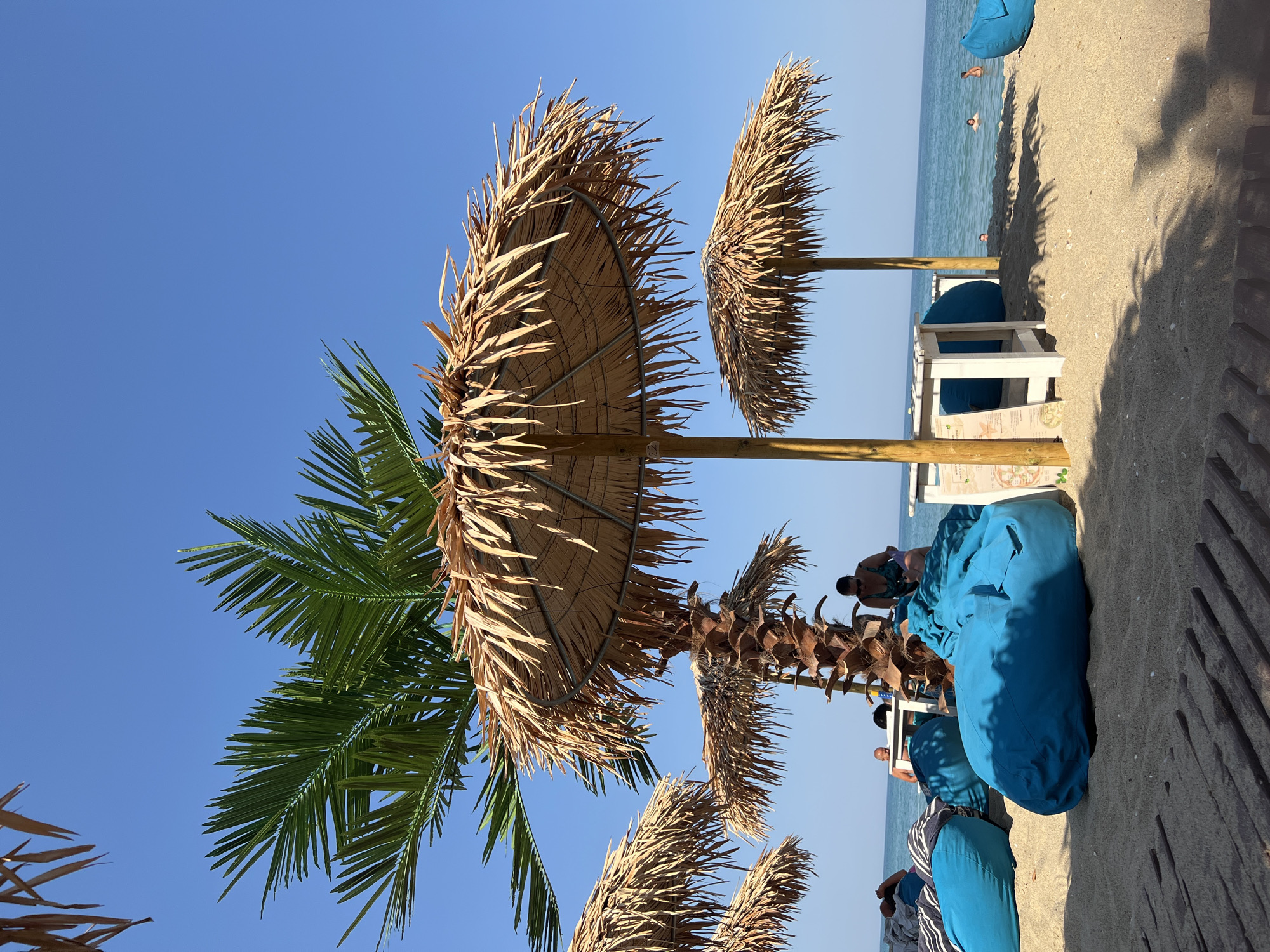 The image depicts a beach scene in Obzor, Bulgaria, under a clear blue sky. In the foreground, a sandy beach is laid out with wooden decking. Several large, bright blue beanbag chairs are scattered across the sand, some occupied by people. Two straw-thatched umbrellas, supported by wooden poles, provide shade, with a palm tree growing between them. Behind the umbrellas and beanbags, the calm blue sea stretches to the horizon, with a few people visible swimming or wading in the water. A woman is seated near one of the tables and umbrellas, looking down. The overall atmosphere is relaxed and summery.