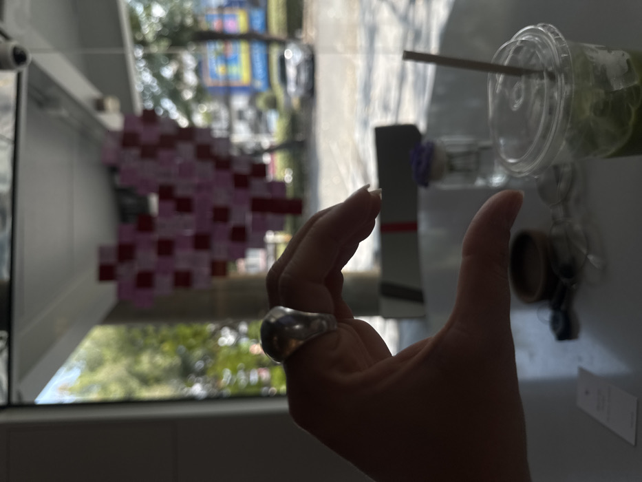 A hand with a large silver ring is in the foreground. In the background, visible through a window, is an abstract pixelated art piece in pink and red hues and greenery. A clear plastic cup containing a green beverage with a straw sits on a white surface, along with a small, dark-colored container and what appears to be a folded card. A reflective surface adds to the complexity of the composition.