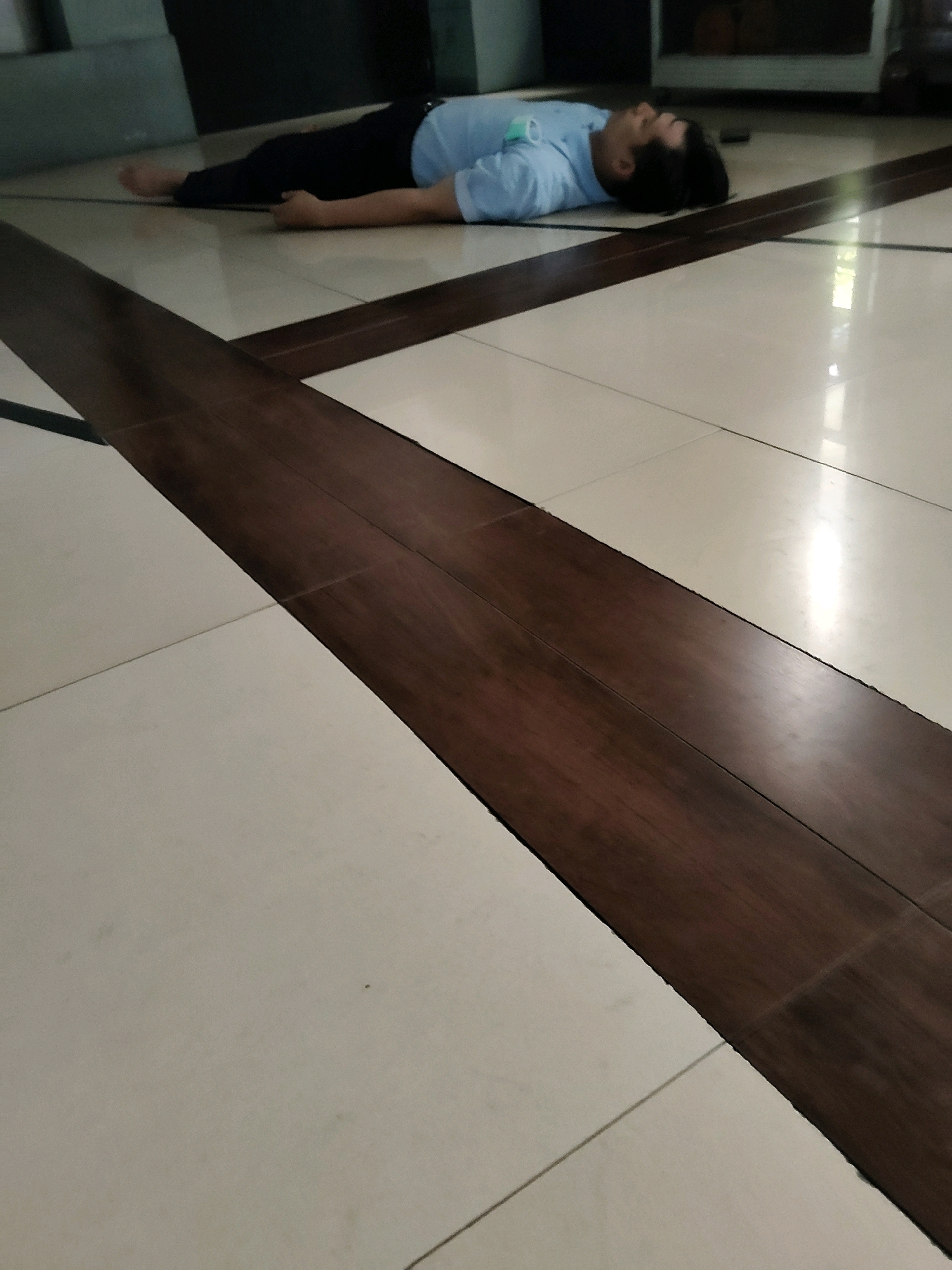 A man in a light blue short-sleeved shirt and dark pants is lying on his back on a tiled floor. His eyes are closed, and his head is turned to his right. The floor is made of large, square, cream-colored tiles, with a diagonal inlay of dark brown wood-look planks running across the image. The room appears to be a public space, possibly a lobby or hallway, with a dark wall and cabinetry visible in the background. The lighting is bright, reflecting off the polished tiles.