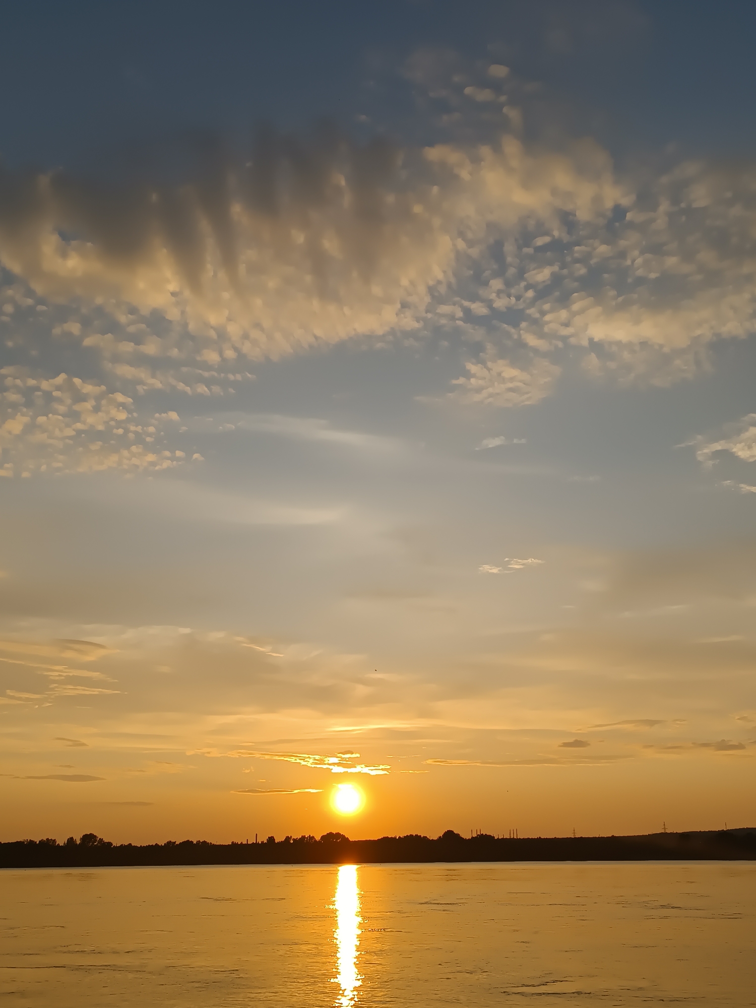 The image depicts a serene sunset over the Kama River in Chaikovsky, Russia. The sun, a bright orange orb, is low on the horizon, casting a golden glow across the water and illuminating a path of light that stretches towards the viewer. The sky is a gradient of warm oranges, yellows, and blues, adorned with scattered clouds that are tinged with the sunset's hues. Silhouetted against the vibrant sky is the distant tree line of the riverbank, punctuated by industrial structures and power line towers, indicative of the city's presence. The water's surface is calm, reflecting the dramatic colors of the sky. There are no people or animals visible in the scene. The overall impression is one of tranquility and natural beauty, with the man-made elements of Chaikovsky forming a subtle backdrop.