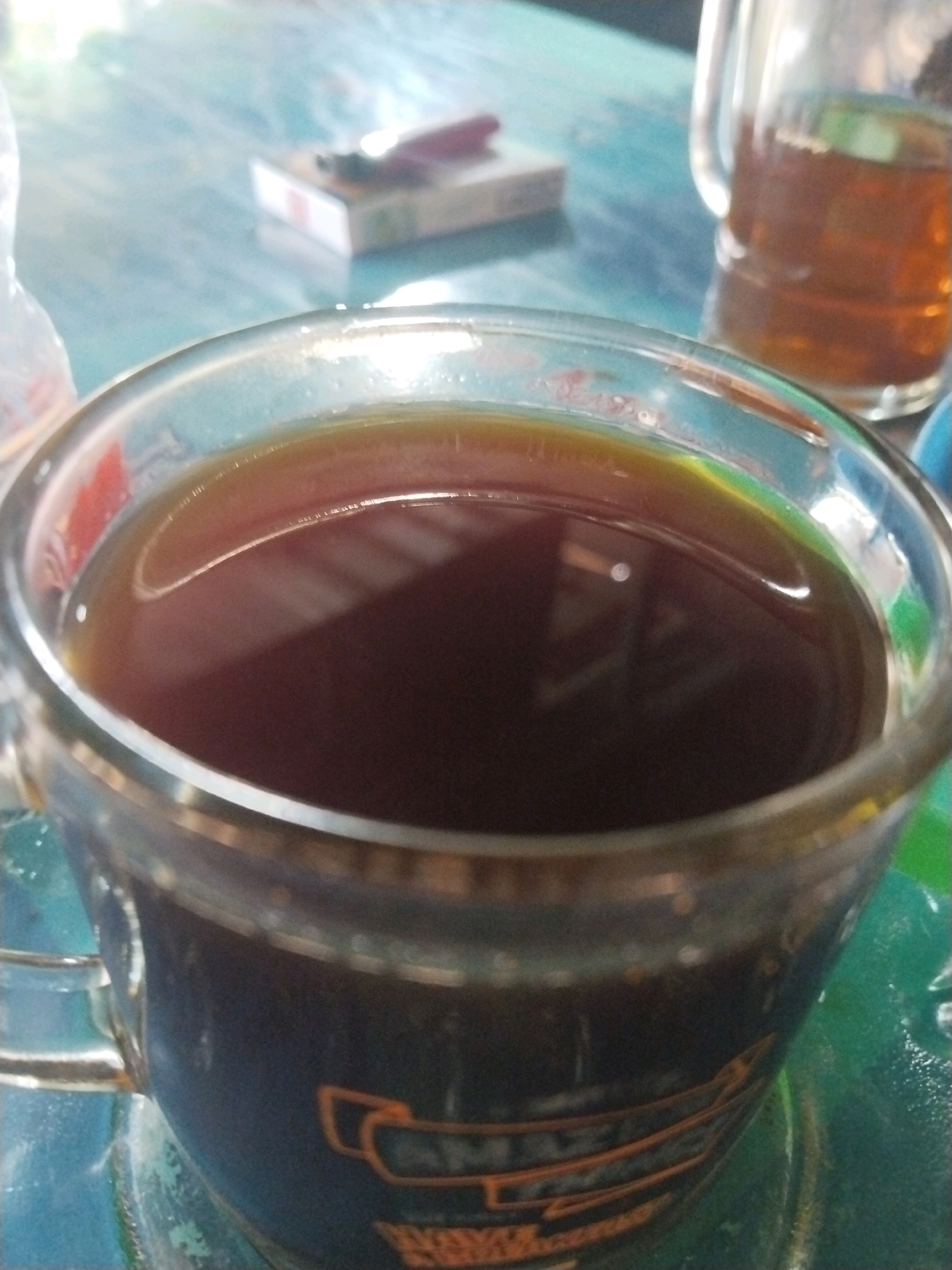 A close-up shot focuses on a glass mug filled with dark coffee, positioned on a teal-colored surface that appears to be a table. The mug has an orange design near its base. In the background, a glass pitcher also containing a dark beverage is visible, along with a pack of cigarettes on the table. The lighting suggests an indoor setting, possibly a cafe or a casual dining area, in Yogyakarta, Indonesia. The overall scene depicts a moment of relaxation or casual consumption.