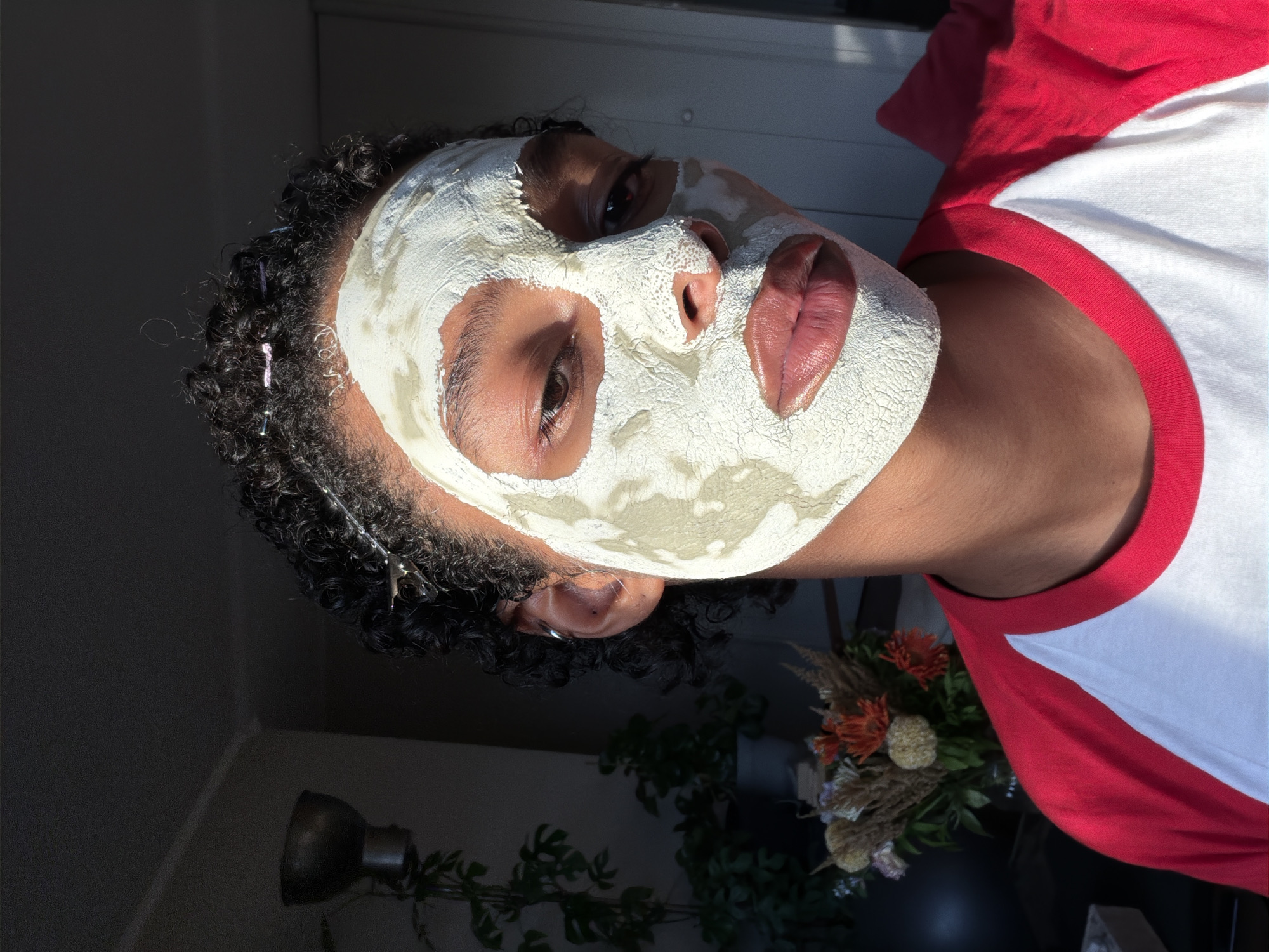A close-up, eye-level shot shows a person with curly dark hair and a white clay mask covering their face. The mask is unevenly applied, revealing the person's eyes, nose, and lips. The person is wearing a red and white baseball style t-shirt. The background is dark and out of focus, with hints of indoor plants and furniture. The lighting suggests natural light coming from the side, creating shadows on the face and a warm glow. The image is taken indoors, likely in a residential setting.