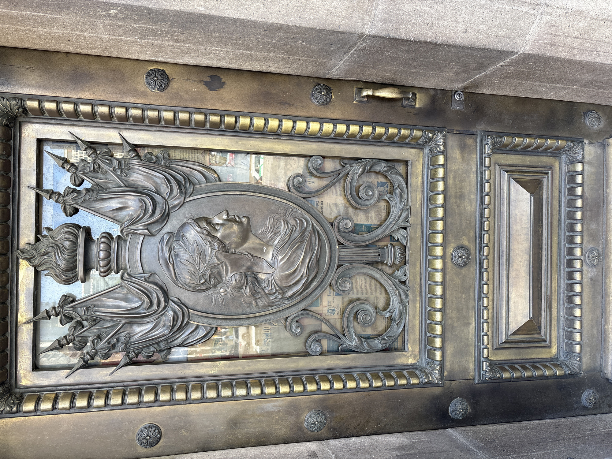 A close-up, frontal view depicts a tall, ornate bronze door, located at the Banco de México building on Avenida 5 de Mayo #2 in the Centro Histórico of Mexico City, Mexico. The door is adorned with a large, dark bronze relief centered on its upper section. This relief features an oval medallion containing a female profile in bas-relief, facing right, wearing a laurel wreath. Above the medallion, a flaming torch emblem rises, flanked by two pairs of draped flags or banners mounted on pointed staffs. Below the medallion, elaborate scrolled motifs complete the central design. These bronze elements are set over a clear glass panel, through which crumpled papers, possibly newspapers with visible Spanish text, are discernible. The door's frame and lower panel are composed of polished and patinated bronze, featuring multiple raised rectangular and segmented moldings. Decorative bronze rosettes are affixed to the door's vertical edges. On the right side, a simple, curved brass handle and a keyhole are present. The door is recessed slightly within a surrounding light-colored stone or concrete architectural element.