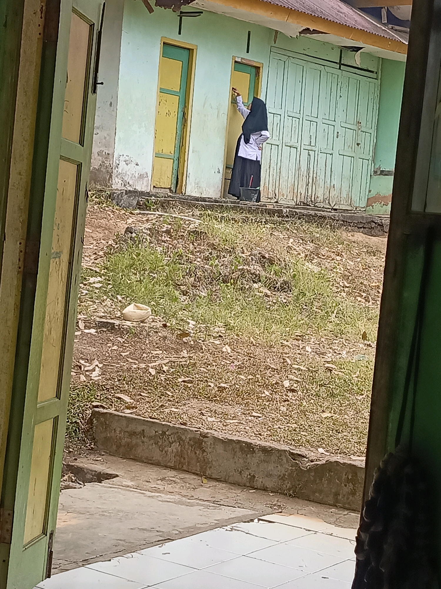 A young girl in a white shirt and black skirt is cleaning a window on the exterior of a light green building. The building has a yellow door next to the window and a large set of light green double doors to the right. The ground in front of the building is covered in dry grass and dirt. There is a concrete planter box in the foreground. The image is framed by an open door on the left and the edge of a dark doorway on the right.
