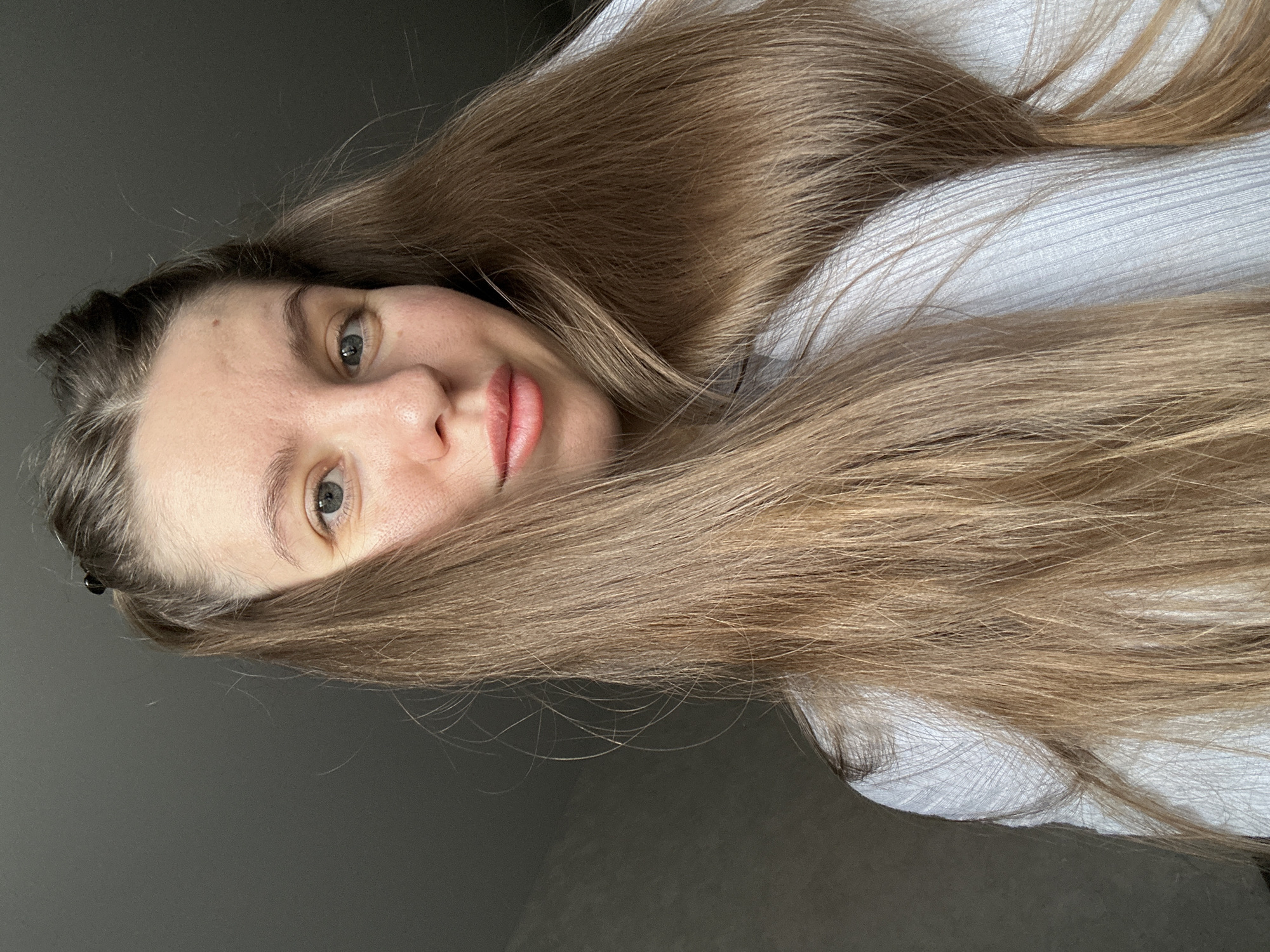 A woman with light brown hair is lying down, her hair partially obscuring her face. She is wearing a white, ribbed sweater. The background is a gray wall. There is no information to confirm location in Klin, Russia.