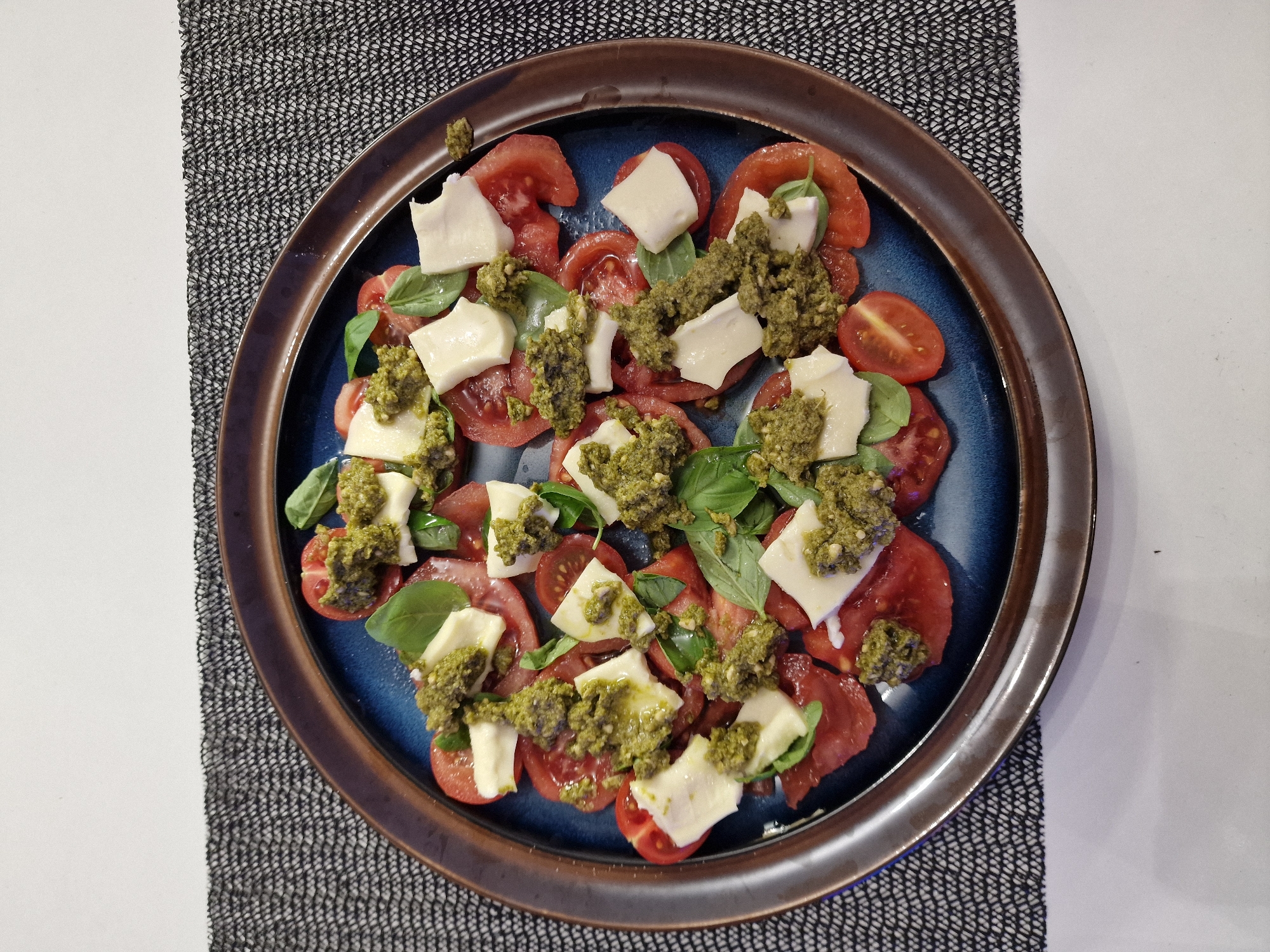 A top-down, eye-level, medium shot depicts a plate of food sitting on a tablecloth. The plate is round and has a dark brown rim surrounding a blue interior. Inside the plate, sliced red tomatoes are interspersed with cubes of white cheese, and fresh basil leaves. A green pesto-like topping is generously dolloped over the ingredients. The tablecloth is a textured grey and white weave. The location of this image is unknown, but has been specified as Sofia, Bulgaria. No people are visible. The overall impression is a simple, fresh salad, likely a variation of the Caprese salad.