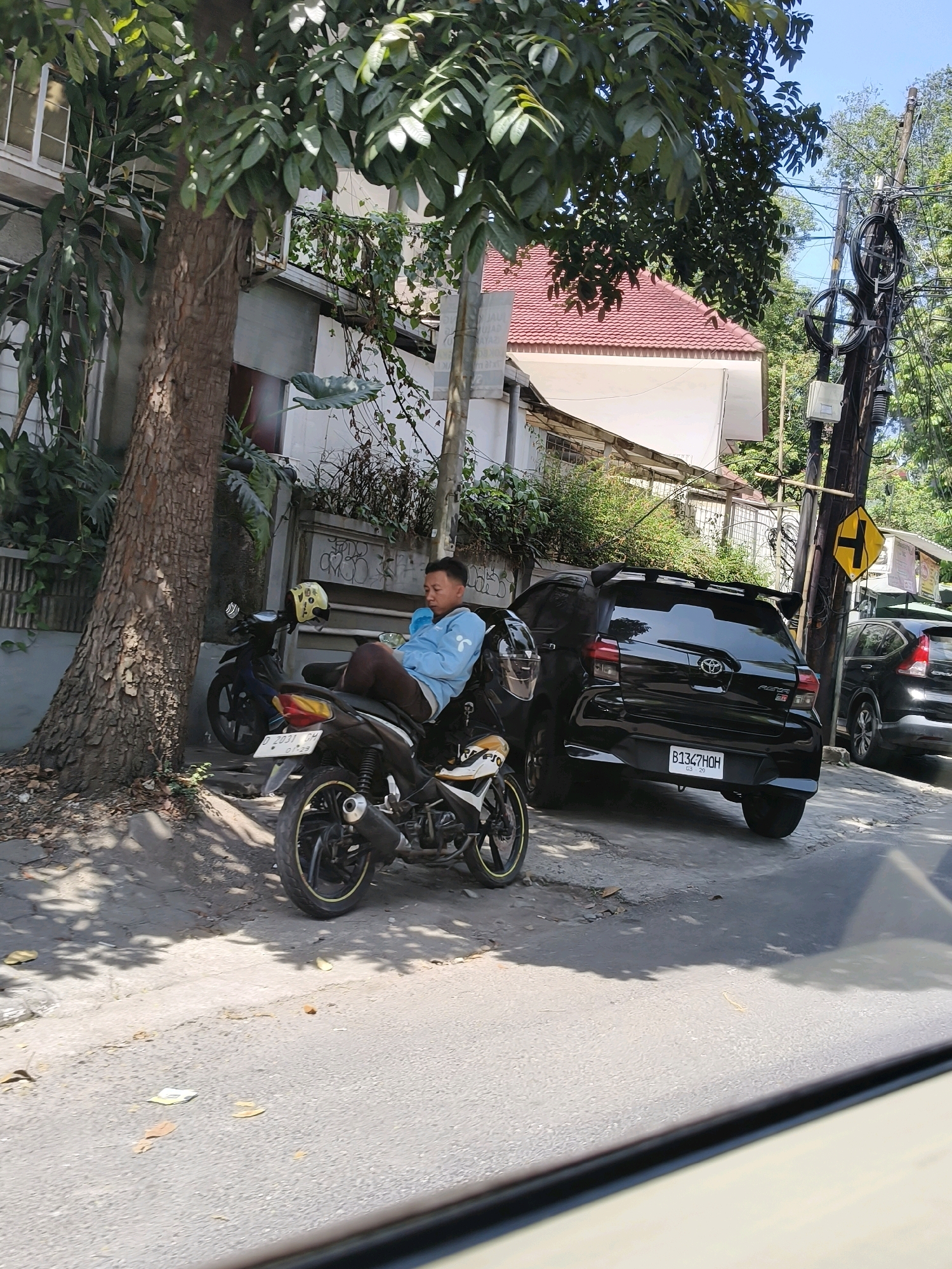 A man is sitting on a motorcycle parked on the side of a street in Bandung City, Indonesia. He is wearing a light blue jacket and dark pants, and is holding a light blue object to his mouth, possibly a drink. The motorcycle is black with yellow accents and has a yellow helmet on the back. A second, darker motorcycle is parked behind it. A black car with a spoiler and a license plate is parked further down the street, along with a black SUV. The street is paved and there is a sidewalk with a tree and some plants. A utility pole with wires and a yellow directional sign is visible on the right side of the image. The buildings in the background have red tile roofs and white walls. The scene appears to be a street in an urban area.