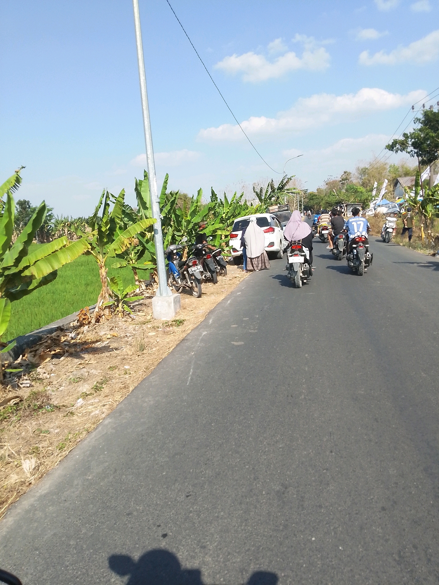 A street scene in Wadang, Indonesia, shows a two-lane asphalt road lined with lush green rice paddies on the left and banana trees on the right. A light pole stands tall on the left side of the road. Several motorcycles and a white car are parked on the right shoulder, and a procession of people on motorcycles is traveling down the road. A person wearing a blue shirt with the number 17 is riding a motorcycle in the middle of the pack, and another person in a pink headscarf is on a motorcycle near the white car. The sky is a clear blue with scattered white clouds, and the overall atmosphere is bright and sunny.