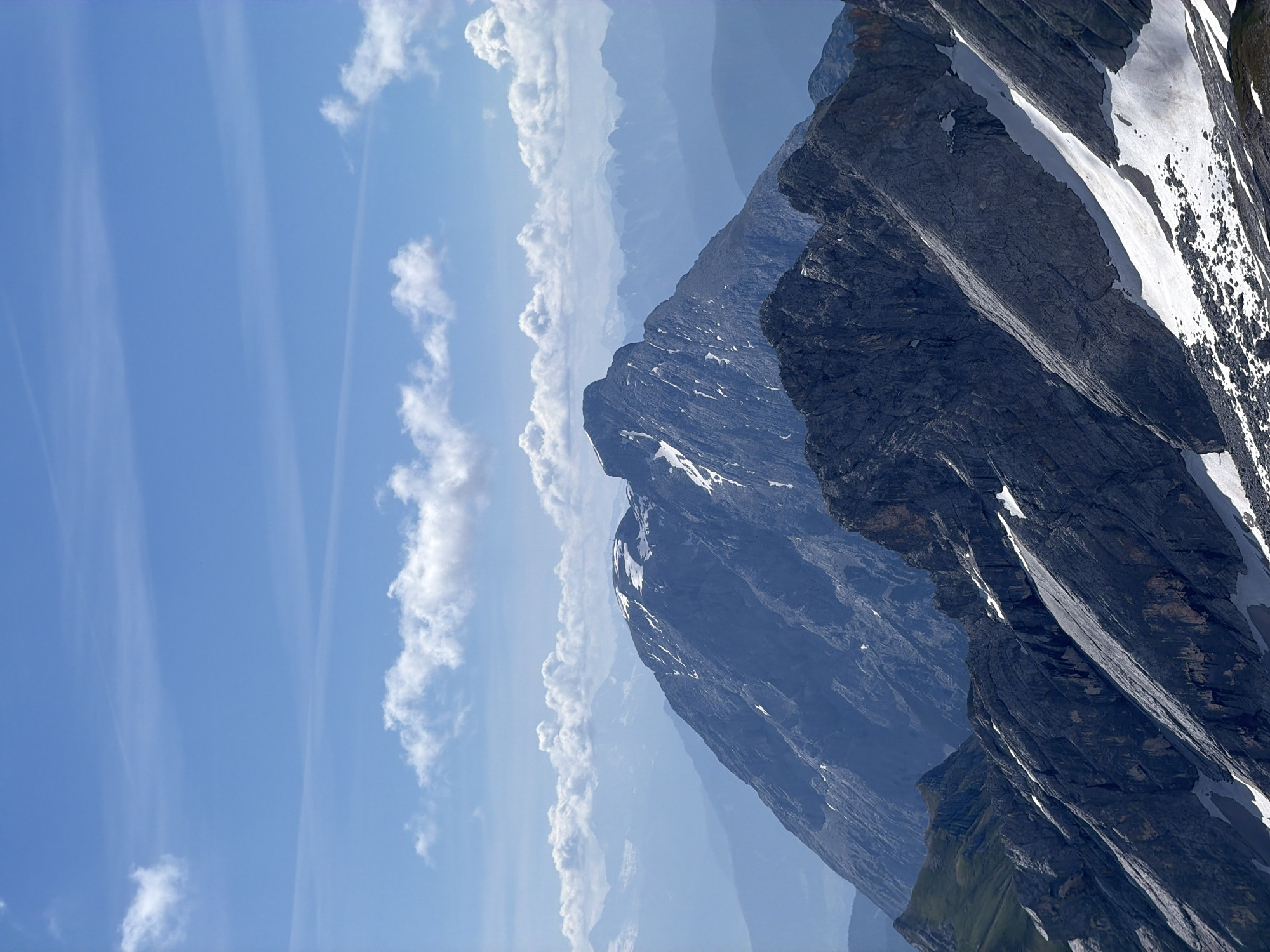This is a vertical, eye-level, outdoor, daytime photograph of a mountain landscape in Garmisch-Partenkirchen, Germany. The image features a towering, rugged mountain range with patches of snow on its slopes. The dominant colors are dark grays of the rocks, white of the snow, and the bright blue of the sky, punctuated by white, wispy clouds.
The mountain face is steep and textured, showing distinct layers and fissures in the rock. The snow appears to be melting in some areas, creating streaks down the mountainside. In the distance, other mountain peaks are visible, fading into a hazy blue, suggesting a vast, expansive landscape. The sky is clear with some thin, elongated clouds that create a sense of depth and movement.
The lighting suggests it is midday, with the sun casting strong shadows on the mountain, highlighting its dramatic contours. The weather appears clear and sunny, typical of a summer or early autumn day in the Alps. There is no activity or event depicted; it is a static shot of the natural scenery. The emotion evoked is one of awe and the grandeur of nature. No text is visible in the image.