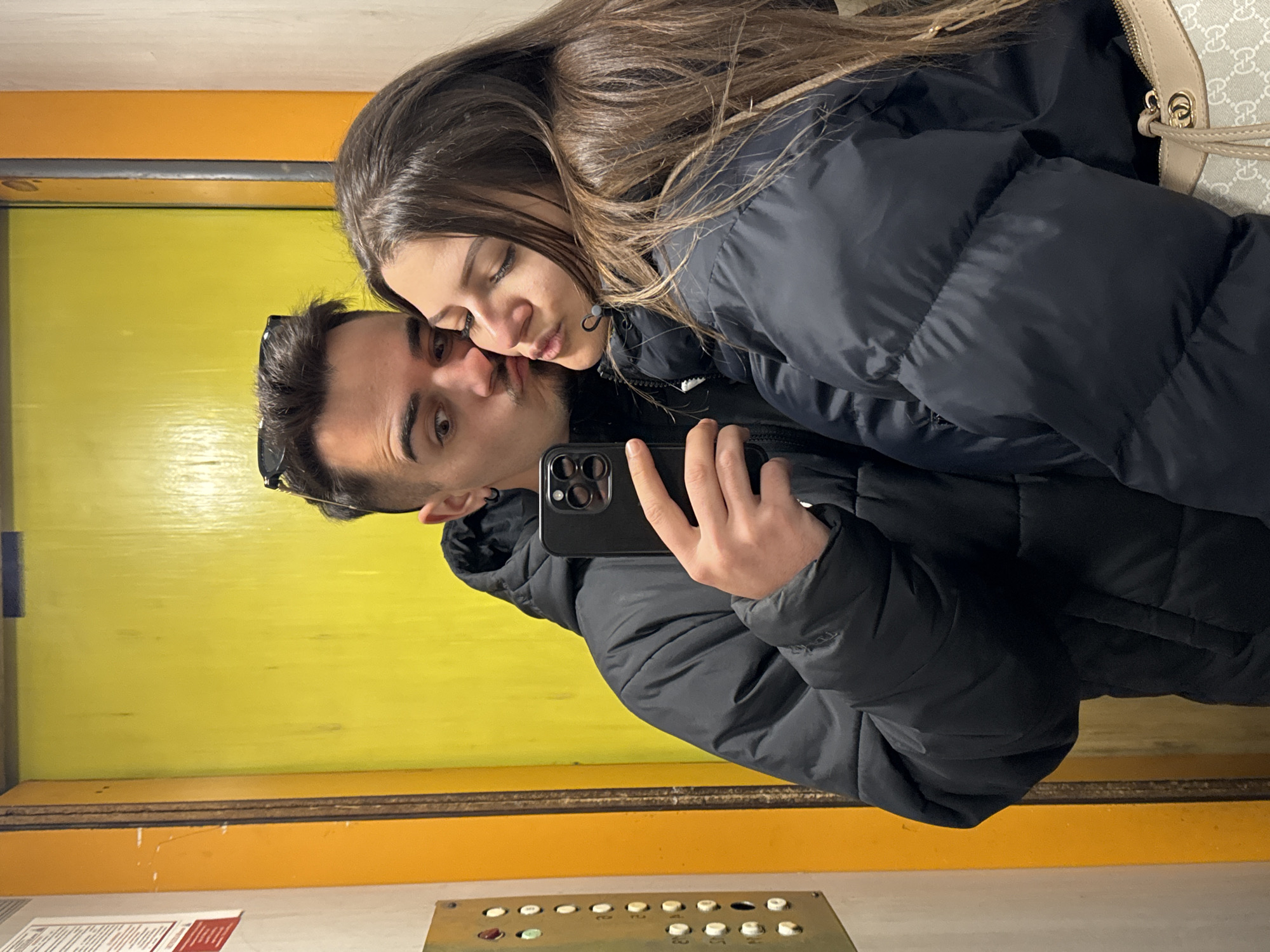 A young man and woman are taking a selfie in an elevator. The elevator has a yellow painted wall with an orange trim, and a vintage elevator button panel with green, red, and off-white buttons. The man is holding a black smartphone and looking at the camera while puckering his lips. The woman has brown hair and her eyes are closed as she leans her face toward the man's cheek. Both are wearing black puffer jackets. A patterned purse is visible on the woman's shoulder. There are red and white documents on the wall. Based on the elevator's appearance and style, this location may be in Sofia, Bulgaria.