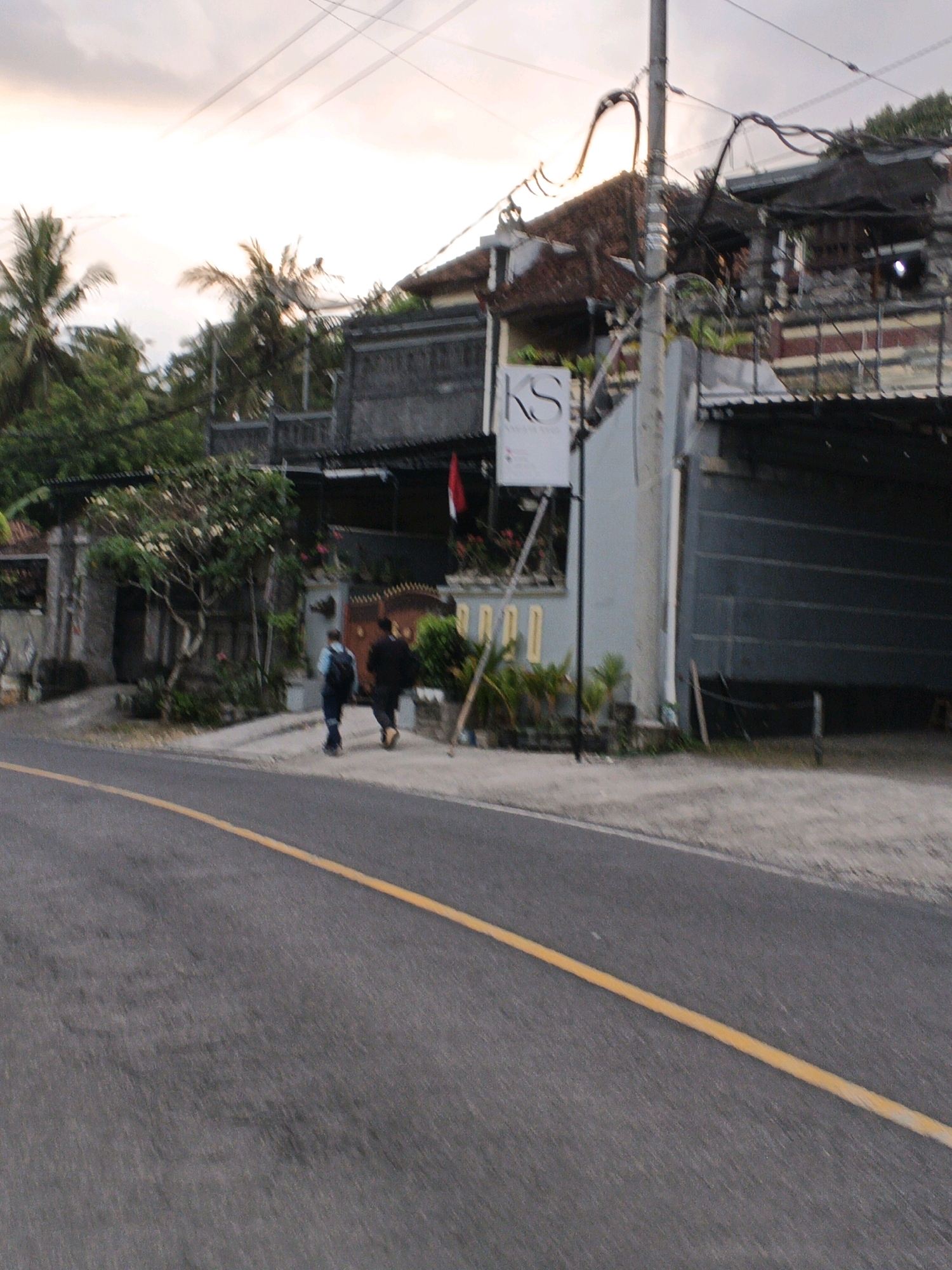 In Manggis, Indonesia, an exterior shot captures a street scene at dusk. Two individuals, one with a blue backpack and the other in dark clothing, walk away from the camera on a paved sidewalk next to a road. A yellow line is painted on the asphalt road. Behind the sidewalk, a building complex features a sign with "KS" and Indonesian text, and a red and white flag. Palm trees and lush vegetation are visible in the background, suggesting a tropical environment. Overhead, power lines crisscross the sky, and the sky is overcast with soft light.
