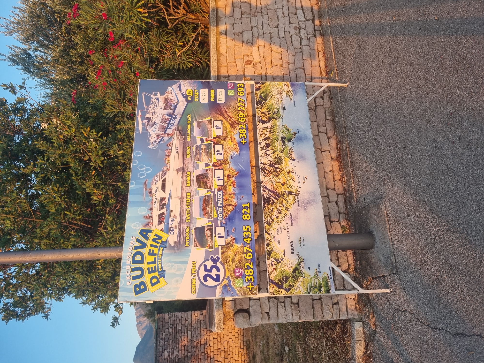 The image shows a large, colorful billboard advertising boat excursions by "BUDVA DELFIN BOATS CRUISE". The billboard is positioned outdoors, in front of a low stone wall and a dense green bush with red flowers. Behind the billboard, a tree with green leaves and a clear blue sky are visible. The billboard itself features images of white boats filled with people, and a map of the Budva Riviera area, indicating various locations like Petrovac, Sveti Stefan, and Kraljičina plaža.
Key information on the billboard includes:
- The name "BUDVA DELFIN BOATS CRUISE" prominently displayed.
- A price of "25€" for excursions.
- Durations for different excursions like "1h" and "2h".
- Contact phone numbers: "+382 67 435 821" and "+382 69 277 693".
- Icons for WhatsApp and a phone call suggest communication methods.
- Details about Wi-Fi, start time (8:10h), and finish time (18:00h).
- A map of the Budva Riviera with place names like Budva, Petrovac, Sveti Stefan, Jaz, and Lastva.
The billboard is supported by metal stands and is situated on an asphalt surface, likely a sidewalk or roadside. The overall impression is an advertisement for tourist activities in a coastal area. Given the map and the prominently featured locations like Budva and Petrovac, it is highly probable that this advertisement is located in or near the city of Bar, Montenegro, as these are popular tourist destinations along the Montenegrin coast. The lighting suggests it is either early morning or late afternoon, with a warm, golden hue cast by the sun.
