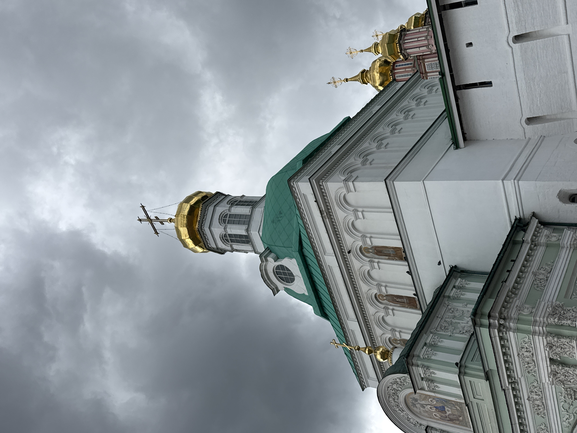 The image shows a low-angle view of the Trinity Lavra of St. Sergius in Sergiev Posad, Russia, under a dramatic, overcast sky. The main focus is on the white and light green facade of the church, adorned with intricate architectural details, including arched windows and decorative moldings. Several golden domes, characteristic of Russian Orthodox churches, are visible, topped with crosses. One prominent dome is a vibrant gold and capped with a cross. Adjacent to it, another section of the church displays multiple smaller gold domes, some with pink accents. The sky is filled with dark, heavy clouds, creating a somber atmosphere. There are no people visible in the frame, and no actions or interactions are depicted. The environment is dominated by the imposing religious architecture.