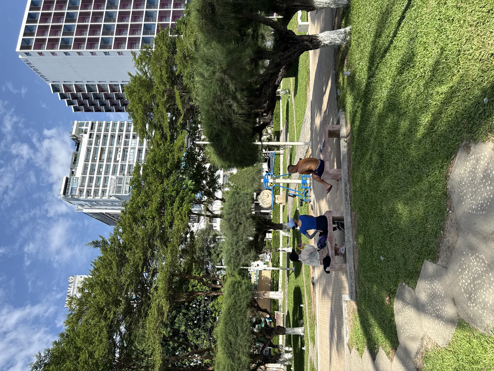 In Nha Trang, Vietnam, a sunny day unfolds in a park adjacent to tall buildings. The scene features lush green grass and trees, some with white-painted trunks, creating a vibrant contrast with the urban backdrop. Three individuals are visible: two women and one man. The women are seated on a stone bench, one holding a phone and the other looking forward. The man sits on a similar bench, his back to the camera, wearing blue shorts. Nearby, outdoor fitness equipment, painted blue and yellow, stands idle. In the background, a cluster of modern buildings, including the Novotel and Panama hotels, pierce the sky. The sky above is a bright blue, dotted with white clouds.
