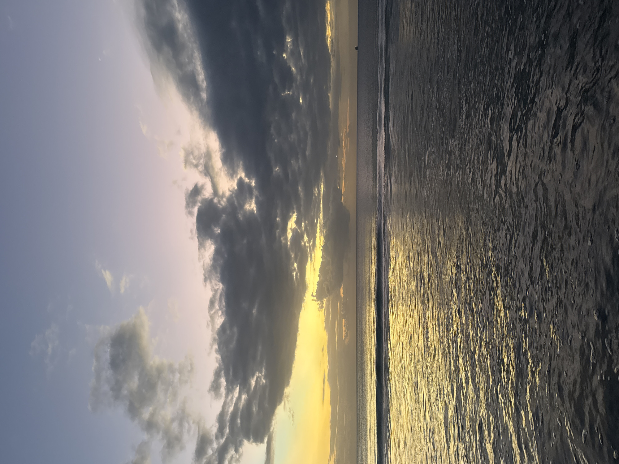 The image depicts a coastal scene, likely at sunset or sunrise, in Kuta Selatan, Indonesia. The sky is partially obscured by dark, heavy clouds with a golden glow from the sun breaking through in the middle, casting a golden color on the water. The ocean extends towards the horizon, exhibiting the motion and texture of waves, with the sunlight reflecting off its surface, creating streaks of light and darkness. The photo seems taken at ground level.