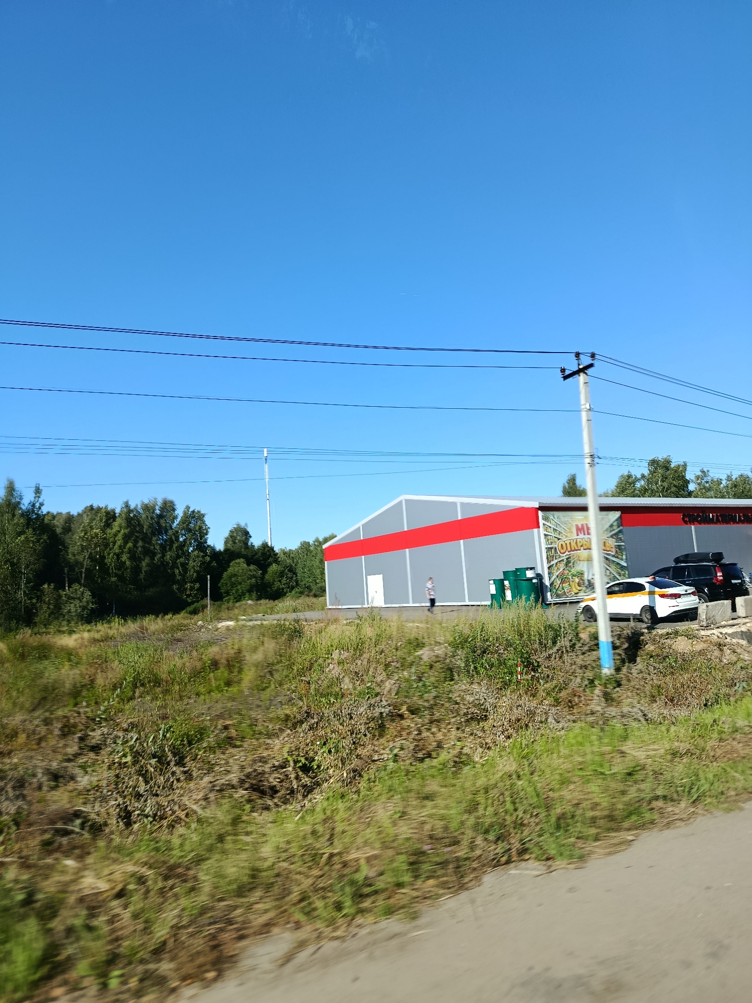 The image shows a bright, sunny day with a clear blue sky above. A modern building with a gray facade and a red stripe runs horizontally across it is visible. The building appears to be a commercial establishment, possibly a supermarket or a warehouse. There is a large advertisement banner on the side of the building that features Cyrillic text and images of food items.
In front of the building, several cars are parked. One car is white with a yellow stripe along its side, and another is a black SUV with a roof rack. A person is walking near the entrance of the building. To the left of the building, there are several green cylindrical objects, possibly barrels or storage containers.
The foreground of the image is dominated by overgrown grass and vegetation, suggesting an undeveloped area adjacent to the building. A power pole with multiple wires is situated in the middle ground, crossing the scene. In the background, a dense line of trees forms a natural boundary. Further in the distance, a tall, slender tower, likely a communication tower or a pylon, stands against the sky. The overall scene appears to be located in a semi-rural or industrial area on the outskirts of a settlement, possibly Snopok Novyi, Russia, given the Cyrillic signage.