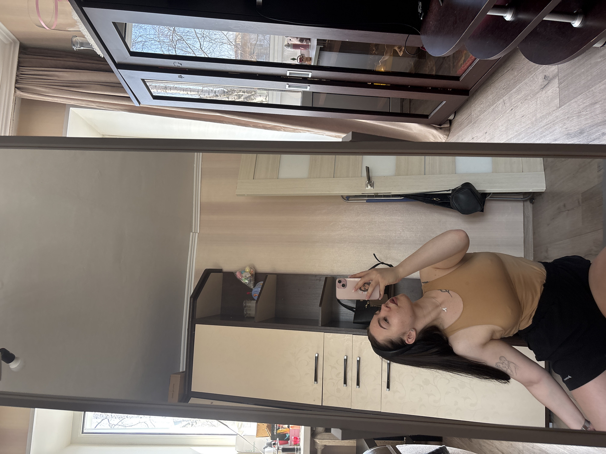 A young female is depicted in a full-length mirror, taking a selfie within an apartment room in Perm, Russia. She is seated or kneeling, angled upwards, wearing a tan sleeveless top and black athletic shorts. Her dark hair is pulled back by a black headband, and she has a silver cross necklace and a tattoo on her upper left arm. She holds a light pink smartphone in her right hand, aimed towards her face, with her gaze directed at the phone's screen.
The surrounding environment, visible in the mirror's reflection, includes light wood laminate flooring and beige wallpapered walls. To the subject's left, a light-toned wardrobe with patterned doors and dark horizontal handles is visible, alongside an integrated shelving unit containing various items. To her right, a dark brown cabinet with glass doors reflects bare tree branches and a bright sky from an adjacent window. A second window is visible in the far left of the reflection. A light wooden door with an opaque glass panel and a hanging black bag are visible in the background, reflected behind the subject. The white ceiling features a single black and white track light fixture. A dark brown tiered shelving unit is partially visible in the bottom right of the reflection.