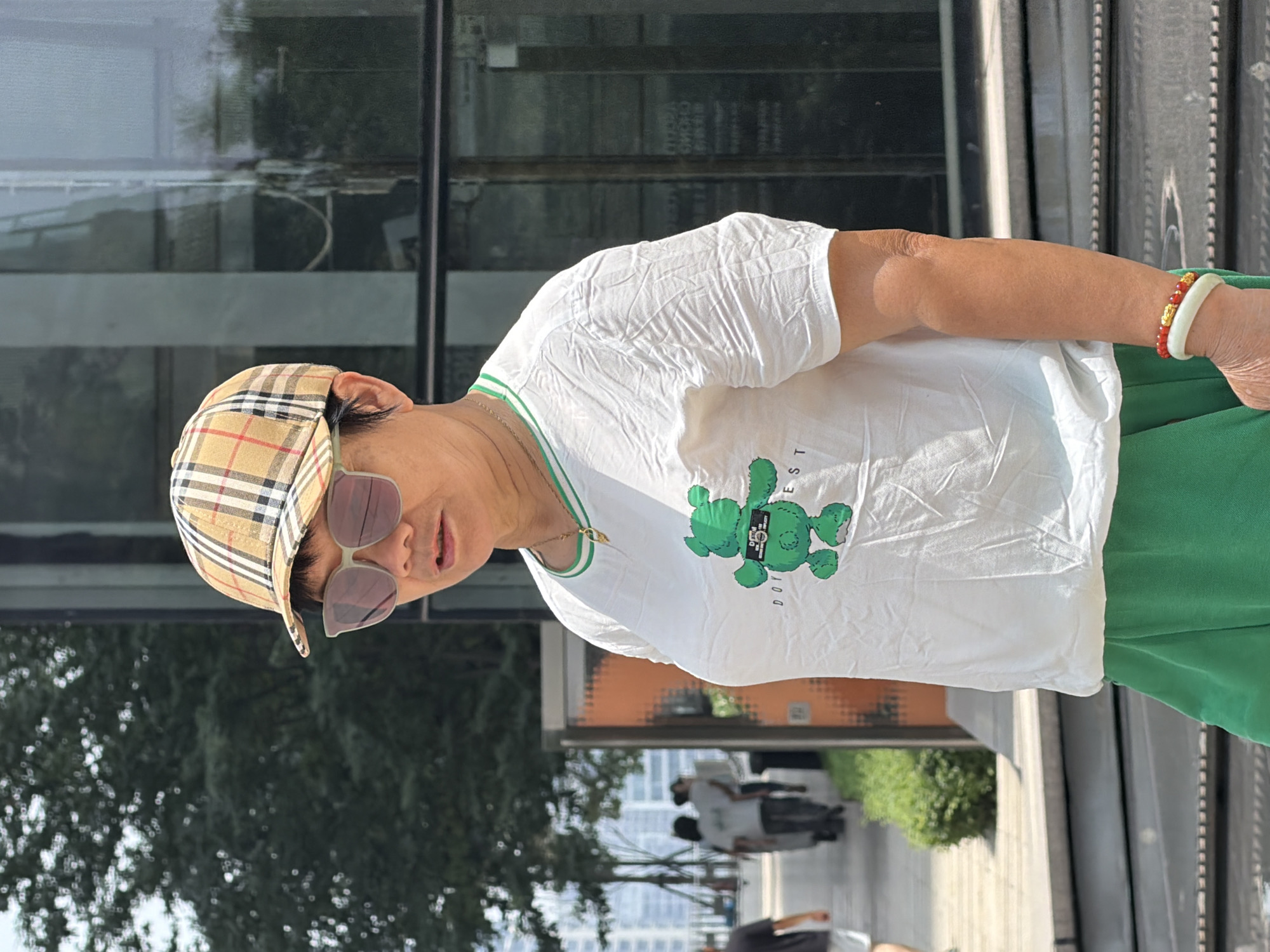 In Yuzhong District, China, an East Asian woman stands outdoors, facing right. She wears a plaid baseball cap and oversized sunglasses. Her white t-shirt features a green cartoon bear graphic and green trim around the neckline. Below her t-shirt, she wears bright green shorts. On her right wrist, she sports two bracelets, one white and one red with gold accents. She appears to be walking on a paved area with some greenery to her left and a modern building with large glass windows behind her. Several other people are visible in the background walking along a pathway. The lighting suggests it is daytime.