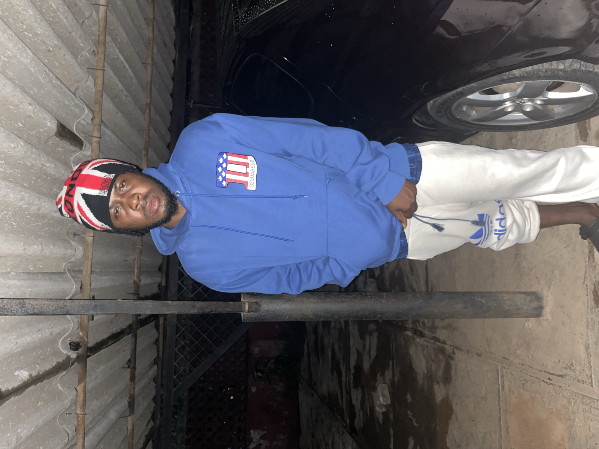 A man stands in front of a black car under a corrugated metal roof. He is wearing a blue hoodie with a graphic on the left chest, white sweatpants with an Adidas logo, and a beanie with the Union Jack pattern. The setting appears to be an outdoor covered area, possibly a parking spot or a workshop, with a concrete floor and a chain-link fence in the background. The lighting suggests it is either nighttime or an indoor, dimly lit space. The overall scene is consistent with an urban environment in Jos, Nigeria.
