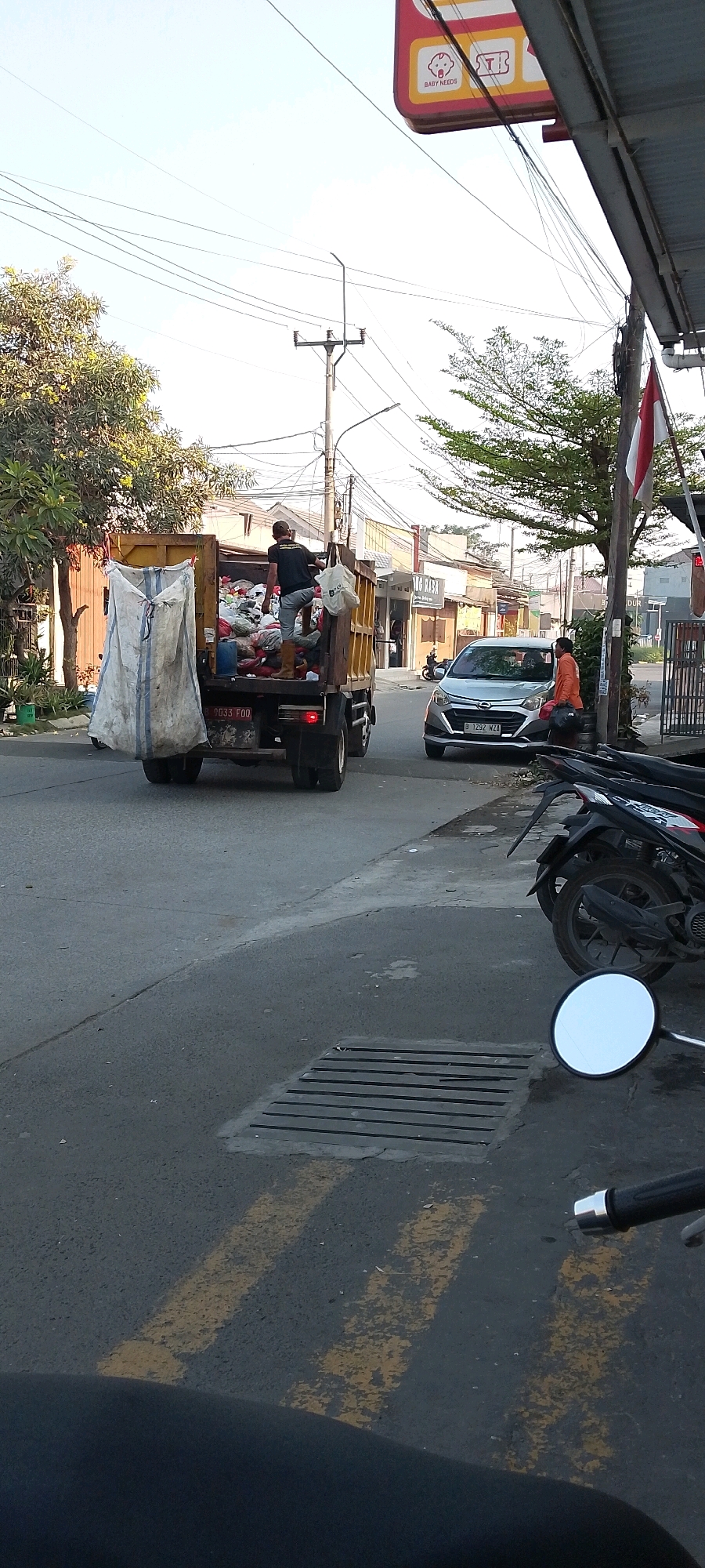 In Satriamekar, Indonesia, a yellow dump truck loaded with white plastic bags and other refuse is parked on the side of a paved road. A person wearing a black t-shirt and brown boots stands on the truck, seemingly sorting or arranging the contents. To the right of the truck, a silver car is stopped, and a person in an orange shirt is standing next to it, holding a helmet. The street is lined with buildings and trees. A partial view of a motorcycle is visible in the foreground, along with a street drain grate. Overhead power lines crisscross the sky. A red and white Indonesian flag is flying from a pole on the right side of the image.