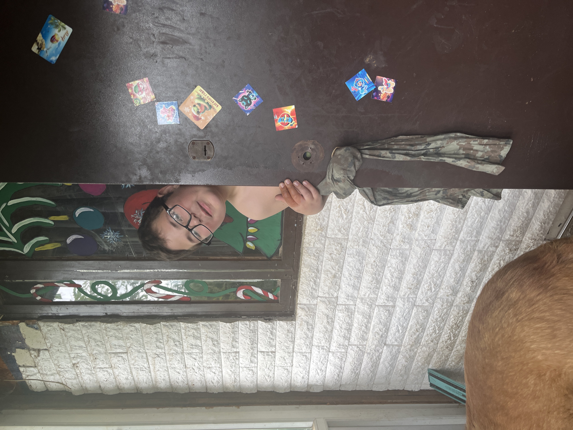 A child wearing glasses is peeking through a gap in a dark brown surface, possibly a door or tabletop, which has several stickers affixed to it. A camouflage-patterned garment is draped next to the child. Below the child, there is a decorated window with festive Christmas-themed designs. A section of textured, white faux stone or brick cladding is below the window. The edge of what appears to be a large dog is visible in the lower right corner. The location, Vesyoloye, Russia, cannot be determined from the visual information provided.