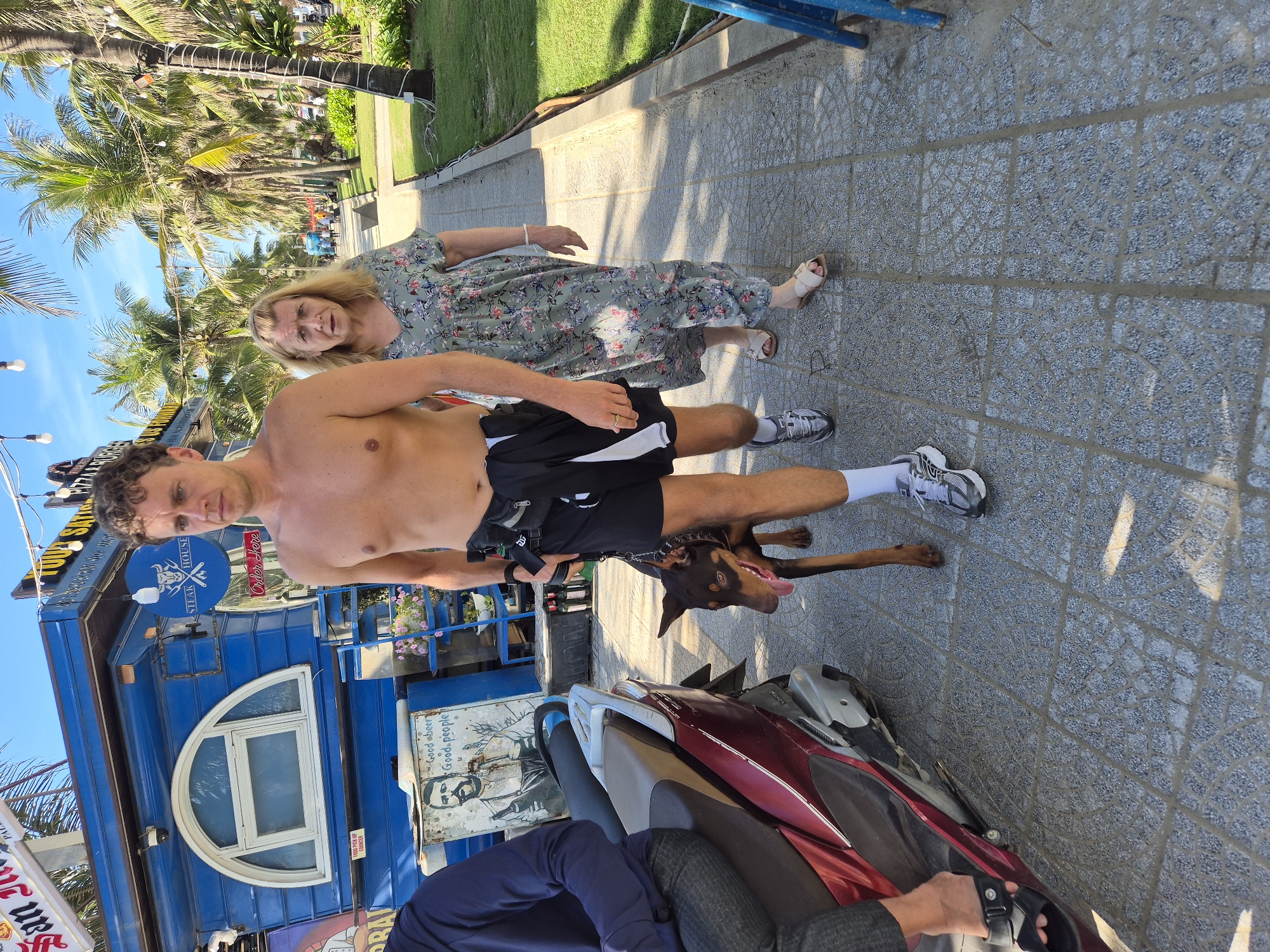 In Đà Nẵng, Vietnam, a shirtless man walks a doberman pinscher on a paved sidewalk. The man is wearing black shorts and grey sneakers. Behind him, a woman in a floral dress walks, her face directed forward. A red scooter is parked to the left of the frame. In the background, a blue building with a arched window is visible. Palm trees and other vegetation line the sidewalk, suggesting a tropical environment. The sky is clear and blue.