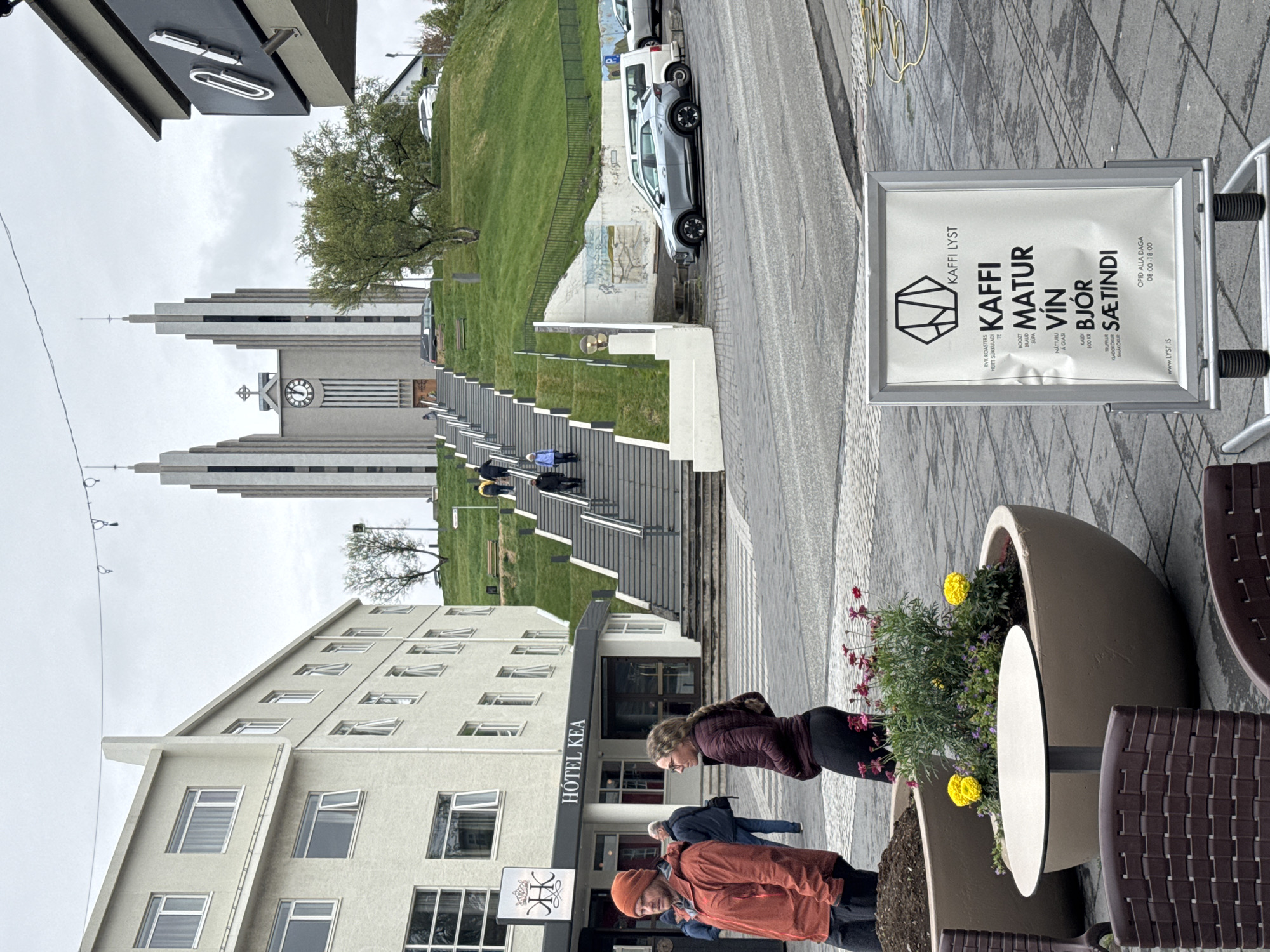 An outdoor street scene in Akureyri, Iceland, is captured on an overcast day. The image, rotated, shows a long, stepped concrete staircase leading up a green hill to Akureyrarkirkja, a prominent modernist church with its distinctive tiered towers and clock face. Several individuals are seen ascending the stairs.
In the foreground, a gray building with multiple windows, identified by the signage "HÓTEL KEA," occupies the left side. To the right, on the sidewalk, outdoor seating comprising dark brown wicker chairs and large planters with yellow and pink flowers create a cafe ambiance. Two people stand near these planters; one woman in a dark jacket looks down, and a man in an orange jacket and beanie looks towards the camera. Another person in a blue jacket is partially visible behind them.
A freestanding sign for "KAFFI LYST" advertises various offerings in Icelandic: "KAFFI" (coffee), "MATUR" (food), "VÍN" (wine), "BJÓR" (beer), and "SÆTINDI" (sweets/pastries). The sign also lists specific items like "RVK ROASTERS HEITT SUKKULADI" (hot chocolate), "BOOZT", "NÁTTURU" (natural), and mentions prices like "600 KR." for "A GLASI" (a glass). It states "OPID ALLA DAGA 08:00-18:00" (Open all days 08:00-18:00) and includes the website "www.LYST.IS".
Beyond the stairs and grassy hill, a street with parked cars, including a silver SUV, is visible. The overall impression is a bustling yet calm urban environment under a cloudy sky.