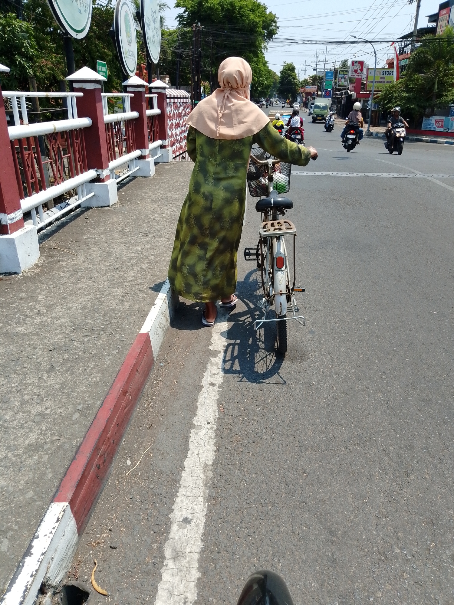 A woman wearing a headscarf and a long green patterned dress walks beside a white bicycle on a sunny day in Pasuruan, Indonesia. The bicycle has a basket in front and a rack on the back. She is walking on a paved sidewalk next to a red and white striped curb. In the background, there are several circular signs with green and white lettering, trees, a street with traffic including motorcycles and a truck, and buildings. The scene appears to be a busy street in a city.