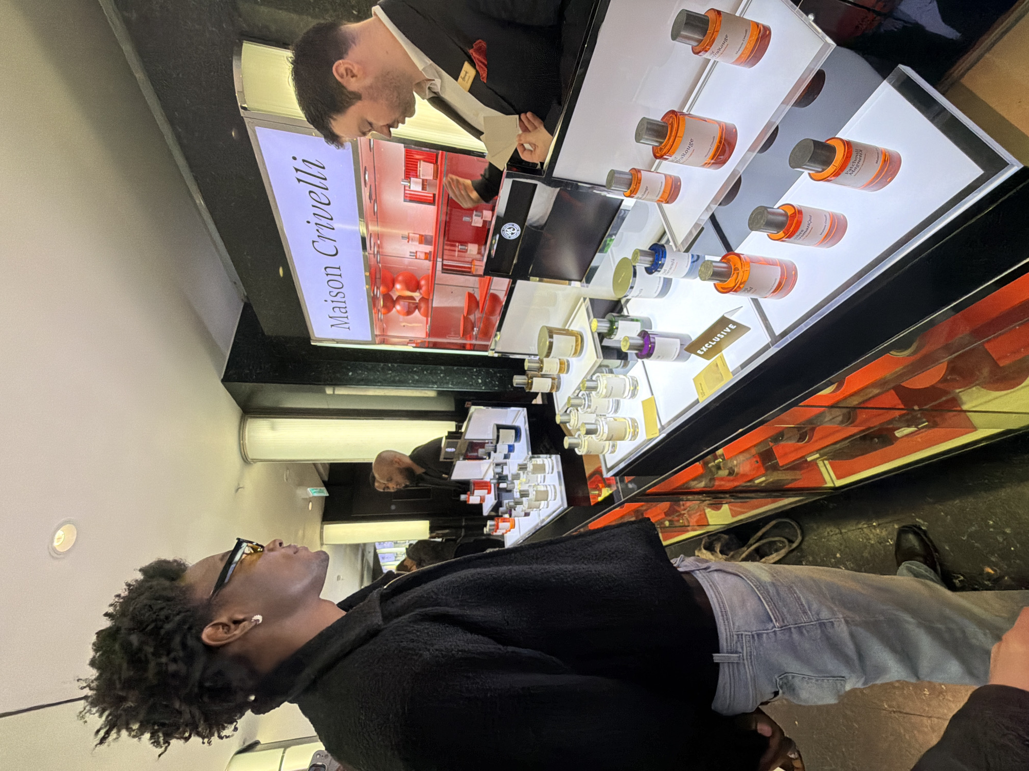 The image depicts an indoor scene at a Maison Crivelli perfume counter, likely located within Harrods department store in Knightsbridge, London, United Kingdom, evidenced by the salesperson's apparent uniform and a logo resembling the Harrods "H" on his name tag.
In the foreground, a Black male, viewed from his right side in profile, stands observing the display and salesperson. He has dark, curly hair, yellow-tinted eyeglasses, and a silver stud earring in his left ear. He is dressed in a dark, textured, zippered jacket and light-wash blue jeans.
To his right, a White male salesperson, seen from his left side in profile, is partially obscured by the display. He has short, dark hair and a beard, wearing a dark suit jacket, white shirt, and a red pocket square. He is holding two white blotter strips, indicative of fragrance sampling, and looking down at the products.
The central subject is a brightly lit, multi-tiered display counter featuring numerous glass perfume bottles from "Maison Crivelli". The bottles contain liquids of various colors, including clear, light amber, orange, purple, and blue, and are capped with dark grey stoppers. Some visible product names include "Citrus Bati", "Patchouli Magnétiq", "Lys Malandia", and "Rose Rouge". A small golden sign marked "EXCLUSIVE" is placed on one shelf. Above this, a backlit sign displays the brand name "Maison Crivelli". Behind the salesperson, a red-lit shelving unit showcases additional bottles and decorative red spheres. The lower portion of the counter features a red interior visible through glass panels.
The background reveals a white ceiling with recessed lighting, dark polished wall surfaces, and reflections of additional blurry figures, including a bald male and another dark-haired male. The floor is dark and reflective. The scene captures a customer interacting with a salesperson at a luxury fragrance boutique.