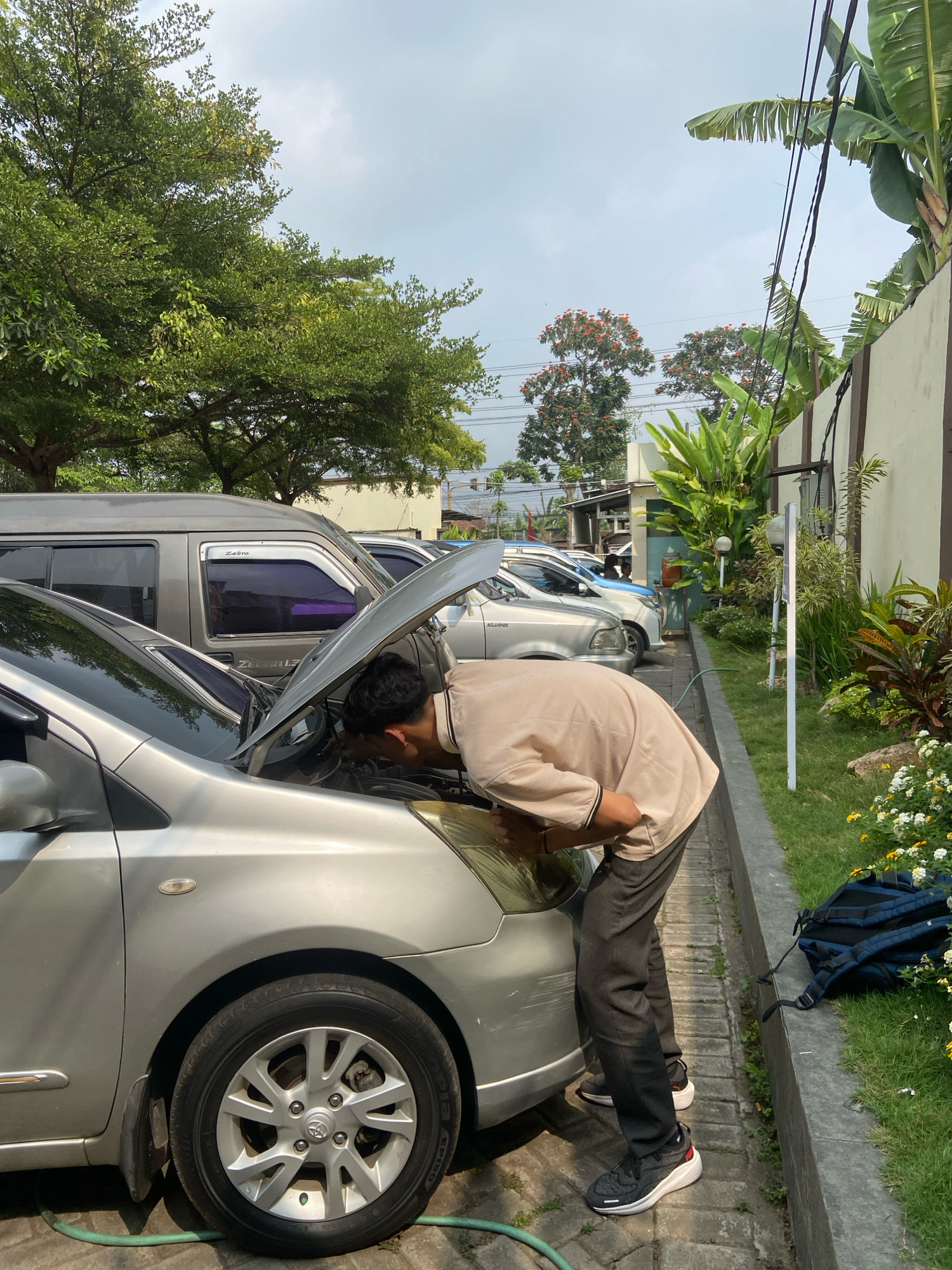 In Kedungwaru, Indonesia, a young man, wearing a light brown collared shirt and dark pants, is inspecting the engine of a silver sedan with its hood open. He is positioned in front of the car, leaning in to examine the engine compartment. The car is parked on a paved surface, possibly a driveway or parking area. To the right of the car, a grassy area is visible with a low concrete border, and beyond that, a wall with some plants. A dark blue backpack rests on the grass near the wall. In the background, several other vehicles are parked, including a gray van. The sky is overcast. The surrounding environment appears to be an outdoor area with trees and vegetation.