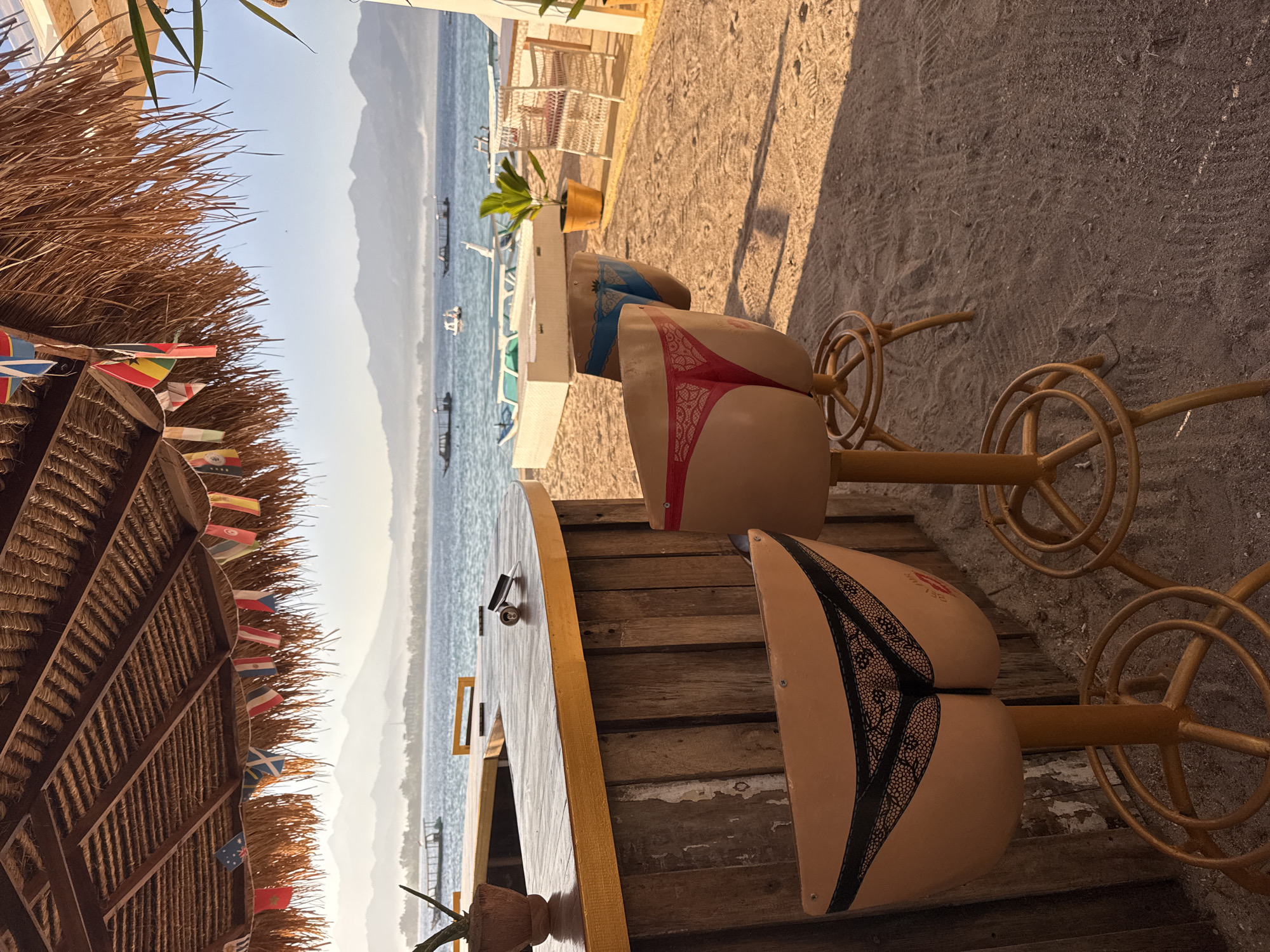 In an outdoor beach bar setting, two novelty bar stools shaped like buttocks wearing thong underwear are positioned in front of a wooden bar. The stools are made of a light-colored material, one with red lace detailing and the other with black lace detailing. Behind the stools, the bar is constructed of weathered vertical wooden planks with a light-colored countertop. A small potted plant sits on the bar. The background shows a body of water with several boats, and a hazy mountainous landscape under a clear sky. The ground is covered in sand. Flags are strung across the thatched roof of the bar area. The scene appears to be located on a beach in an unspecified Indonesian city.