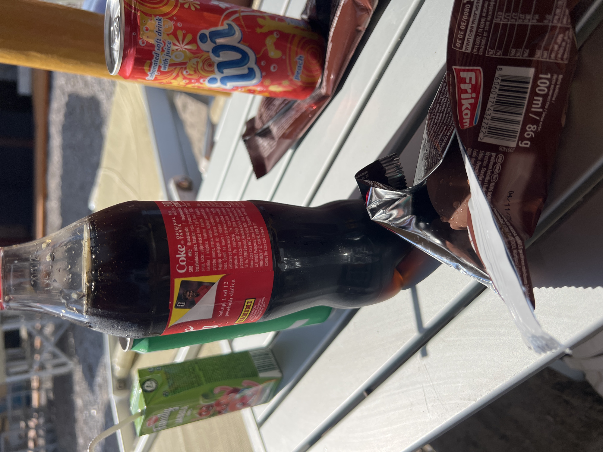 This is a close-up image of a picnic or snack setup on a light-colored, slatted table, likely outdoors. The items include:
* A glass Coca-Cola bottle.
* A can of Ivi peach-flavored carbonated soft drink.
* An open Frikon ice cream bar wrapper.
* A juice box.
The scene appears to be bathed in sunlight, creating bright highlights and shadows. The image seems to have been taken at a slight angle, with the items arranged in a somewhat haphazard manner on the table. There is no indication of the location being in Prijevor, Montenegro.