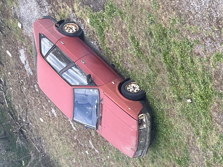 A red, older-model hatchback, consistent with a Moskvitch 2141 "Aleko", is observed parked or stranded on a sloped, uneven terrain in Bolyarovo, Bulgaria. The vehicle is oriented facing downhill, with its front end directed towards the lower part of the slope. The car's paint is faded and appears dirty. The front bumper exhibits damage, with the headlight assembly on the viewer's left (passenger side) missing or severely compromised. The front hood shows a slight misalignment. All four wheels are present, featuring off-white, possibly steel, rims stained with rust and dirt. The surrounding environment consists of patchy green grass intermingled with areas of exposed soil and gravel, indicating a rugged, unpaved surface. Further up the slope in the background, denser vegetation, including trees or shrubs, and scattered dry branches are visible. No individuals or animals are present in the scene, and no actions or interactions are depicted.